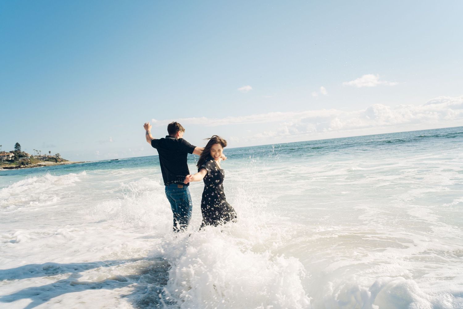Couple splashing in ocean waves at the beach, woman in black dress and man in navy shirt enjoying a romantic moment.