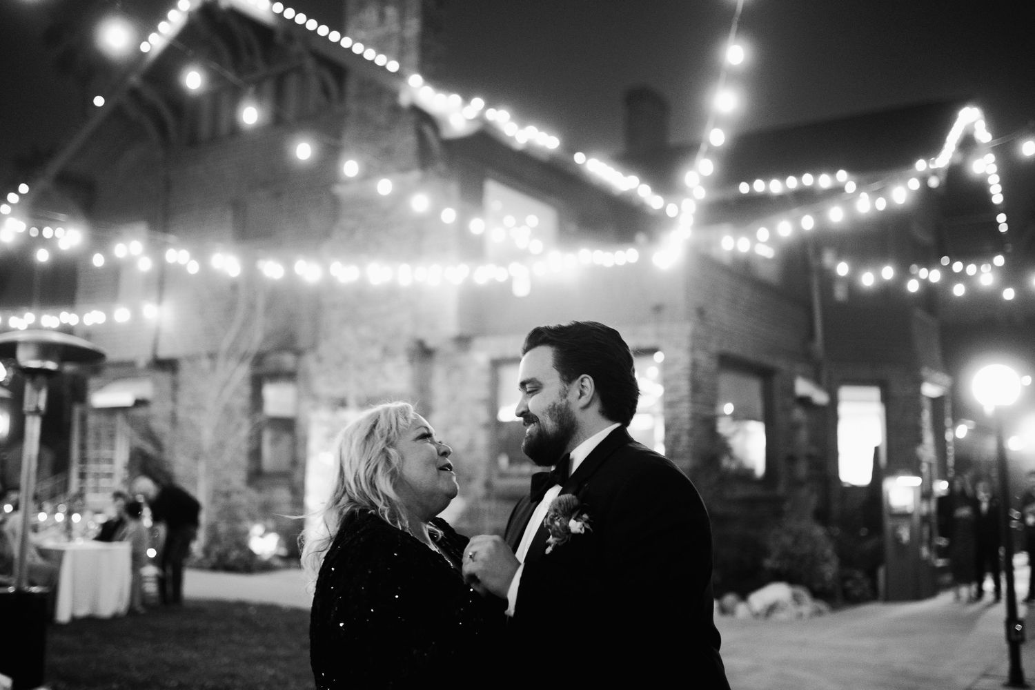 Black and white photo of a couple sharing a romantic moment under string lights at an outdoor wedding reception.