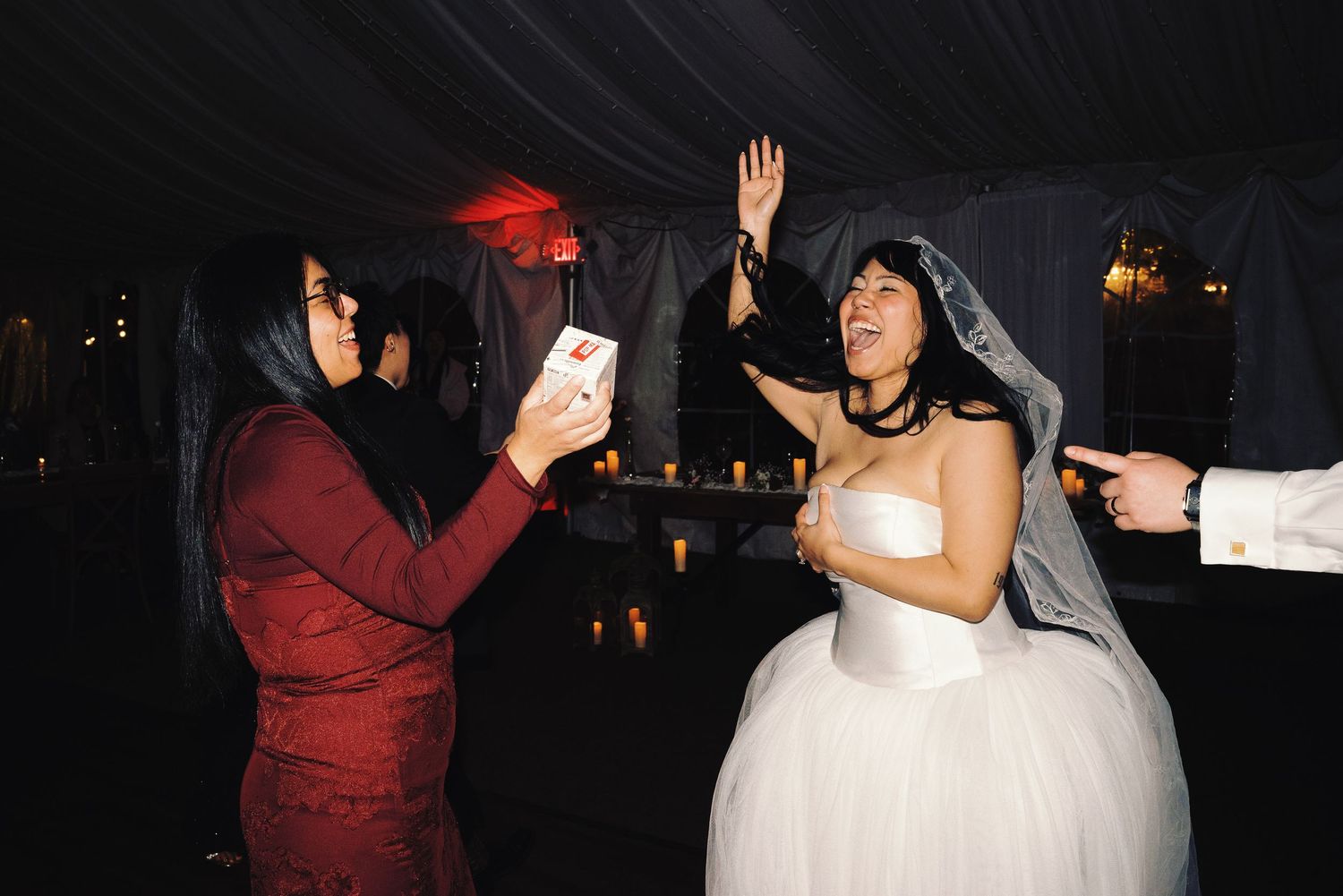 Bride in white gown dancing joyfully while guest captures moment on phone at reception.