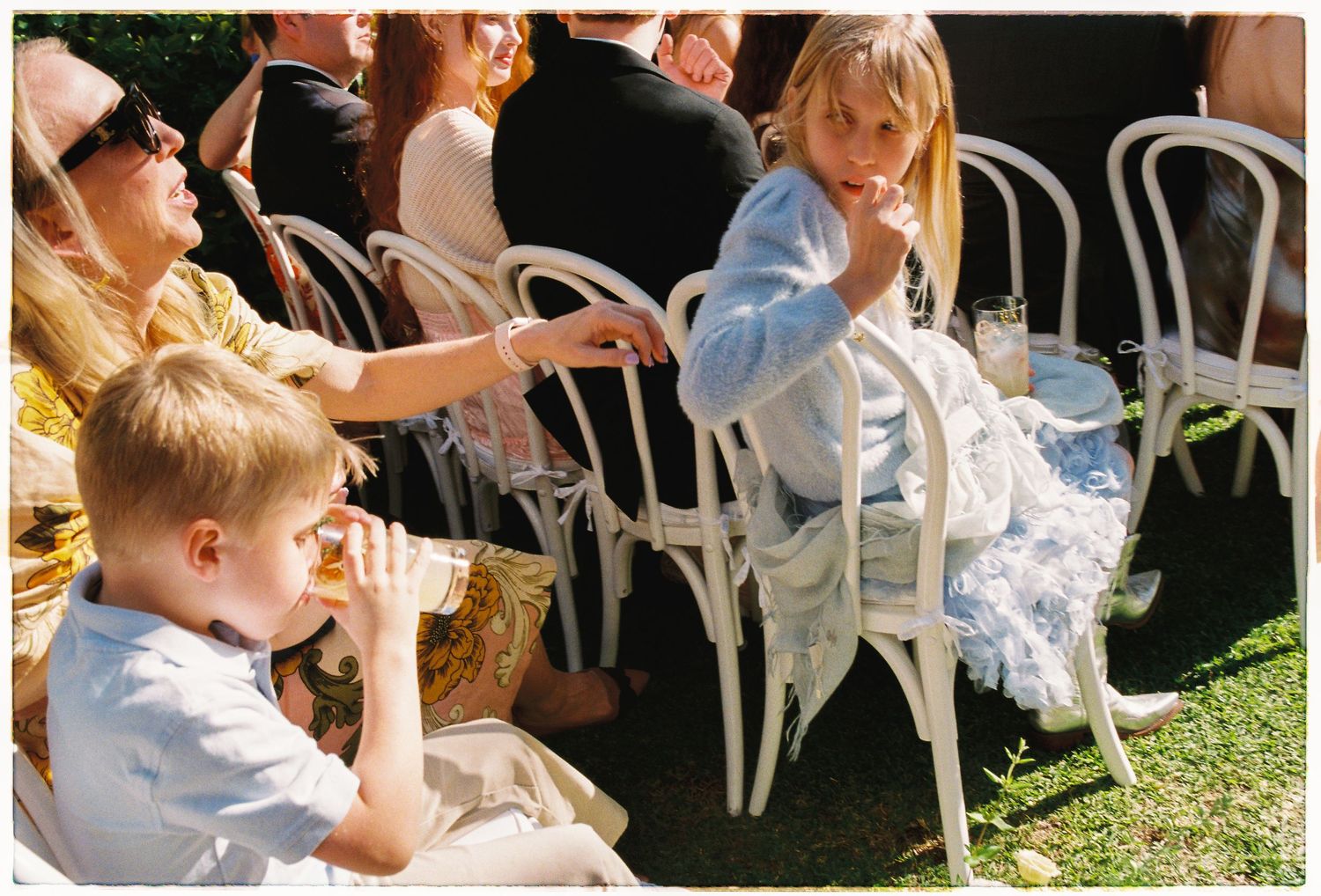 Children in formal attire sit in white chairs at outdoor wedding ceremony on sunny day.