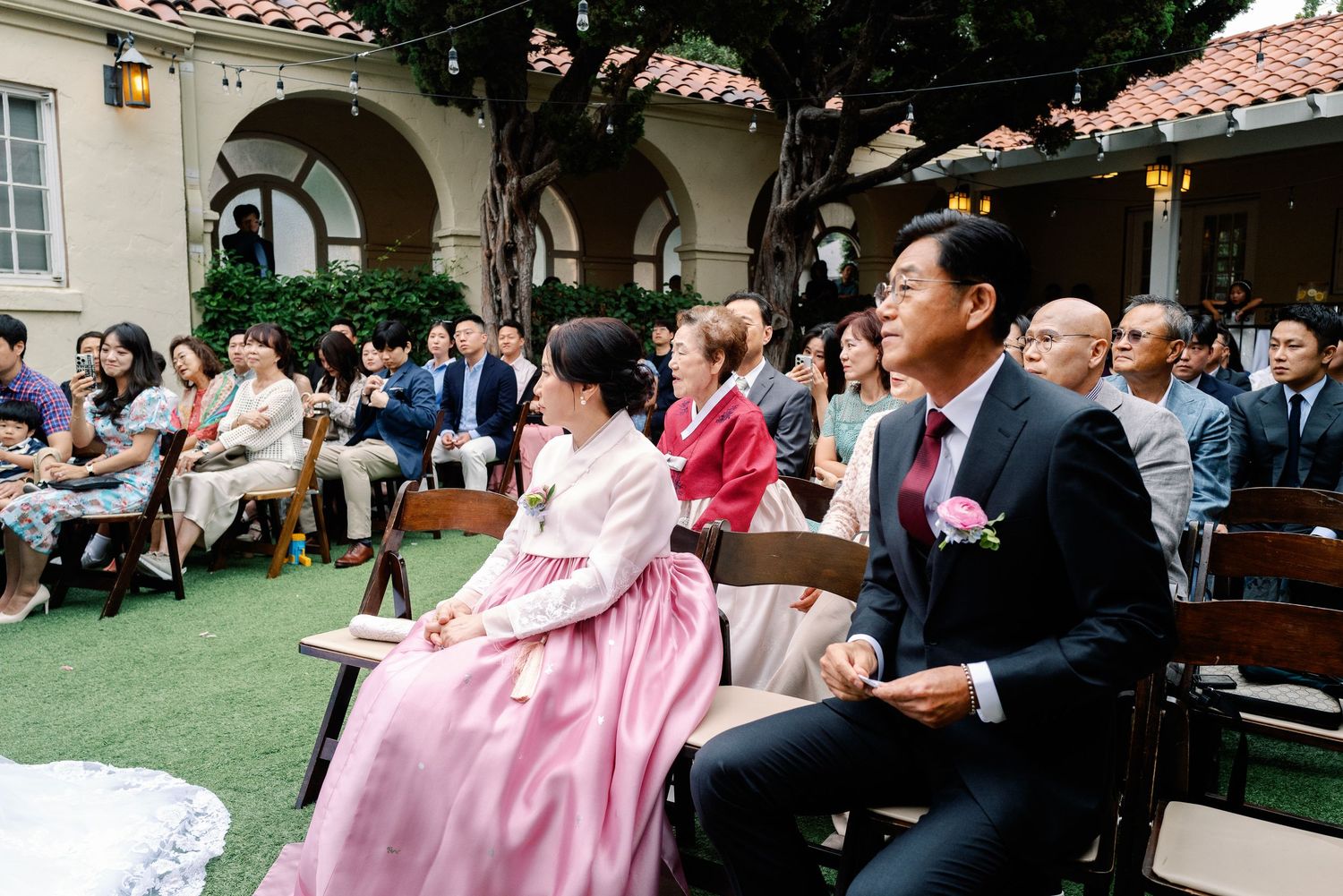 Wedding guests including women in traditional Korean hanbok seated at an outdoor garden ceremony.