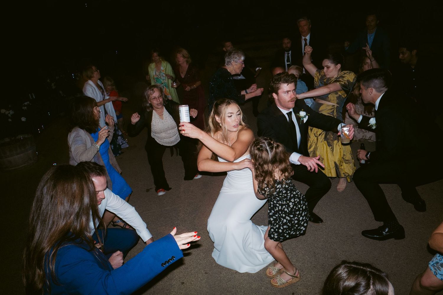 Bride in white gown dancing energetically with guests at lively outdoor wedding reception at night.