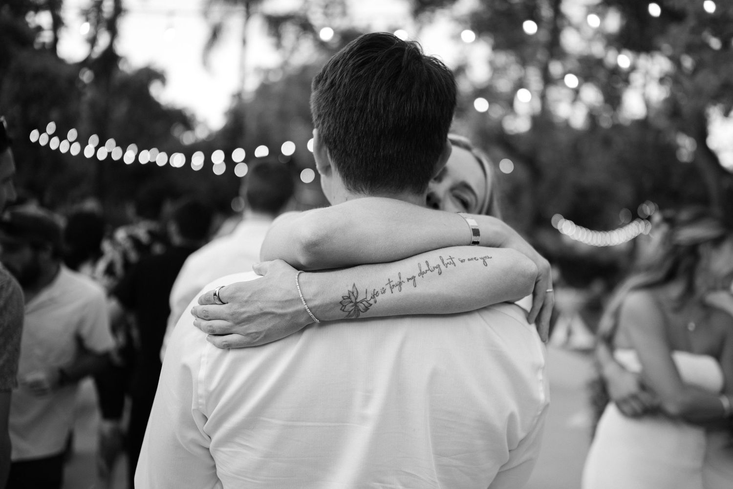 Couple embracing at outdoor wedding reception with string lights, woman showing forearm tattoo.