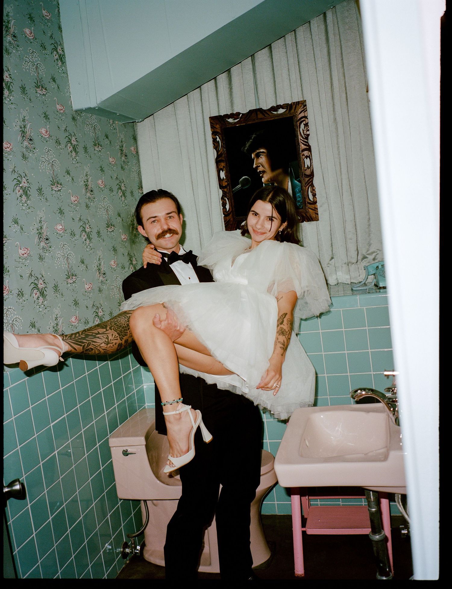 Tattooed groom carrying bride in white dress in vintage pink bathroom with floral wallpaper.