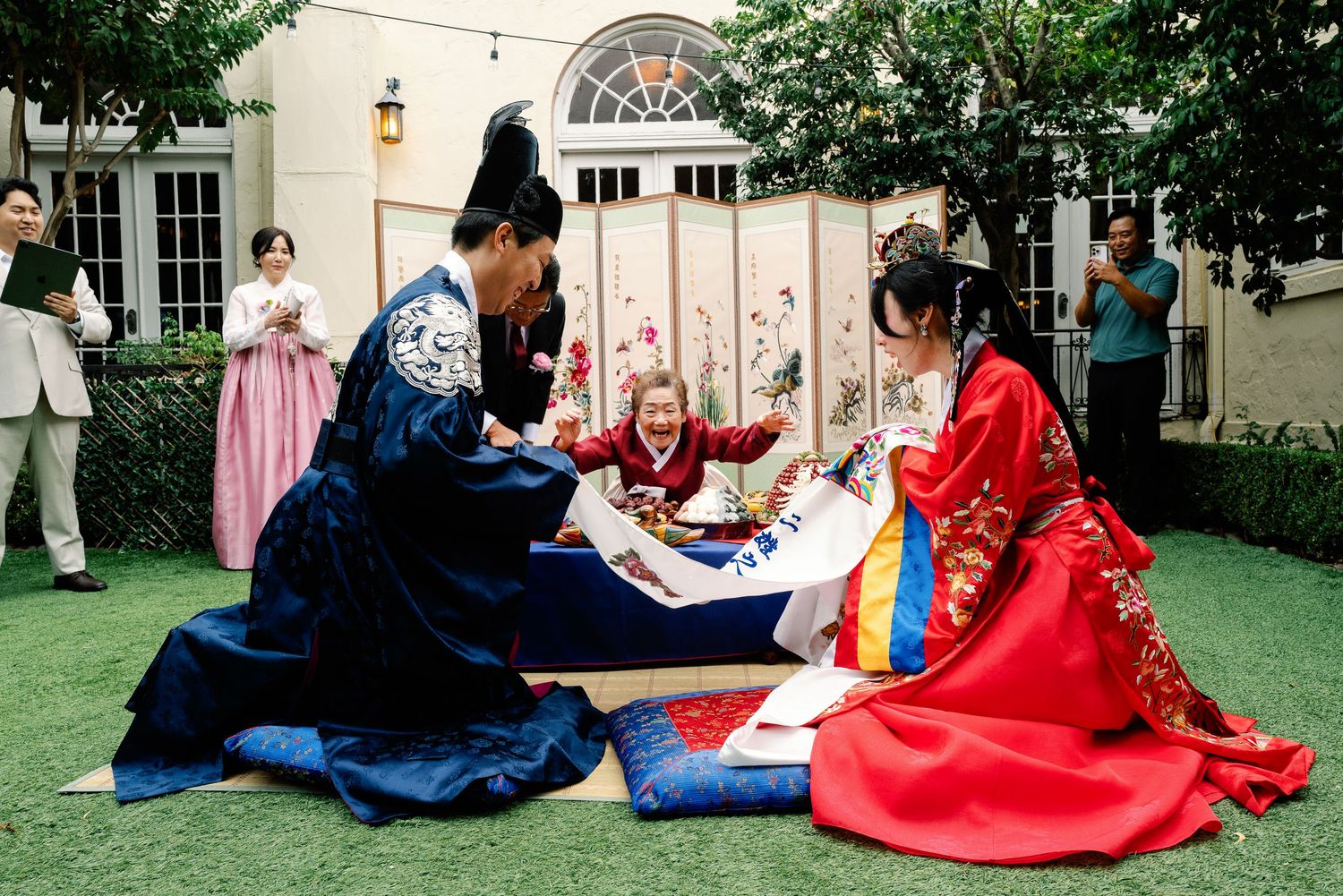 Elderly woman blesses couple in traditional Korean hanbok during outdoor paebaek wedding ceremony.