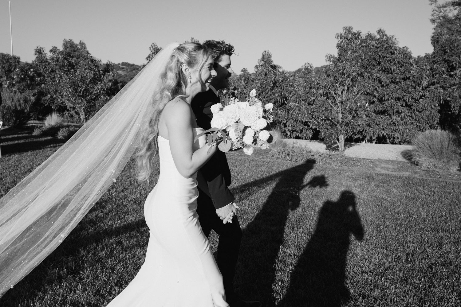 Bride in fitted white gown holding floral bouquet with flowing veil in black and white portrait.