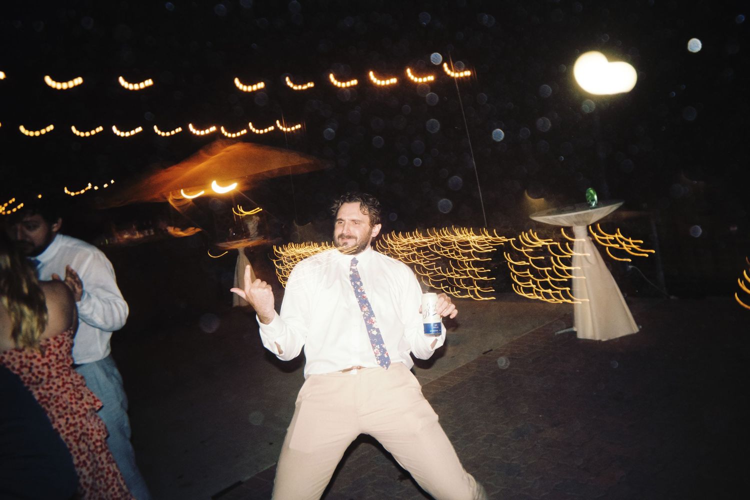 Guest dances energetically on outdoor wedding dance floor under warm string lights at night reception.