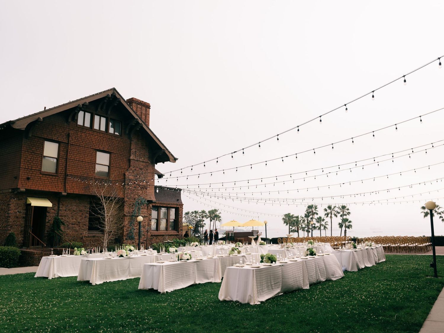 Outdoor wedding reception with white-draped tables on green lawn beside historic brick venue with string lights overhead.
