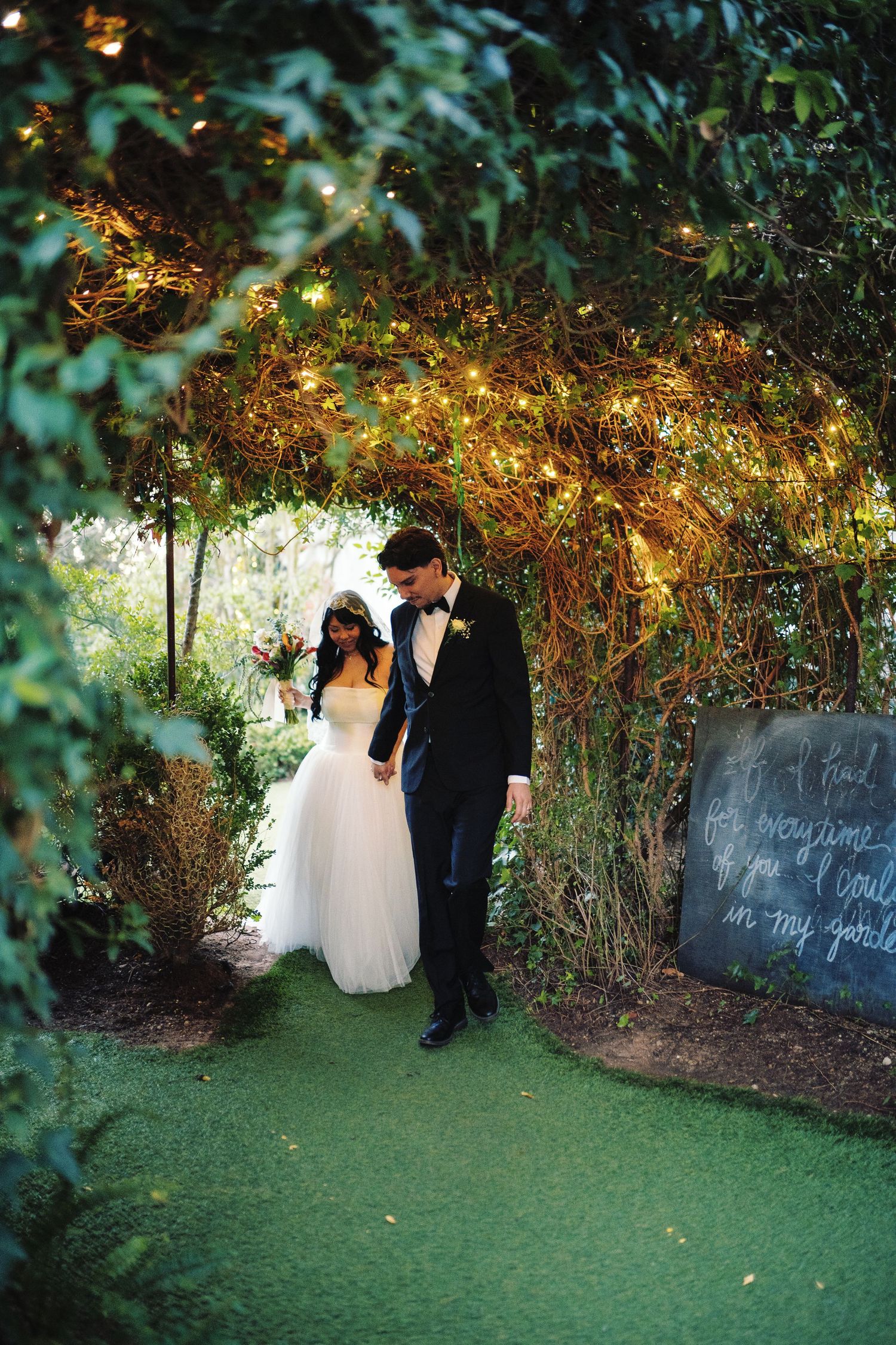 Newlyweds walk hand in hand through a magical lit archway of twinkling fairy lights and greenery.
