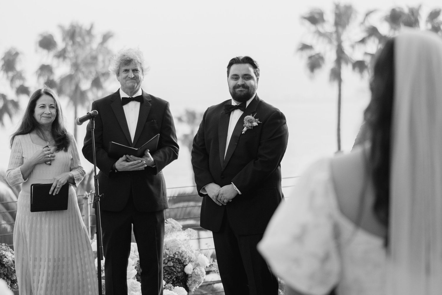 Black and white photo of groom smiling at bride during outdoor wedding ceremony.