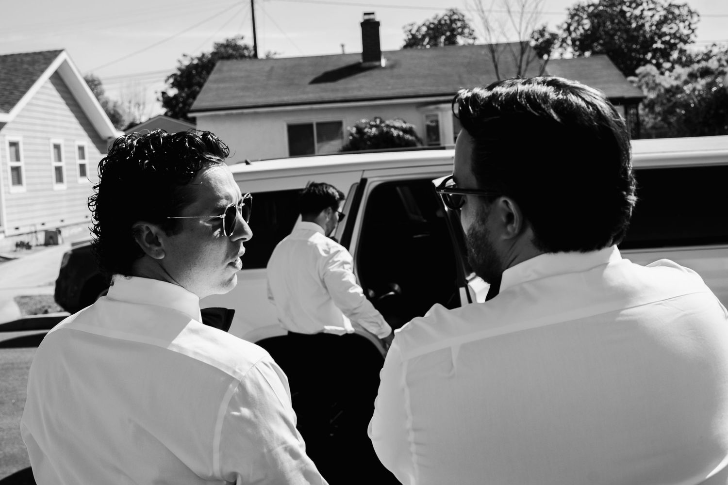 Black and white photo of groomsmen in white shirts gathered near limousine before wedding.