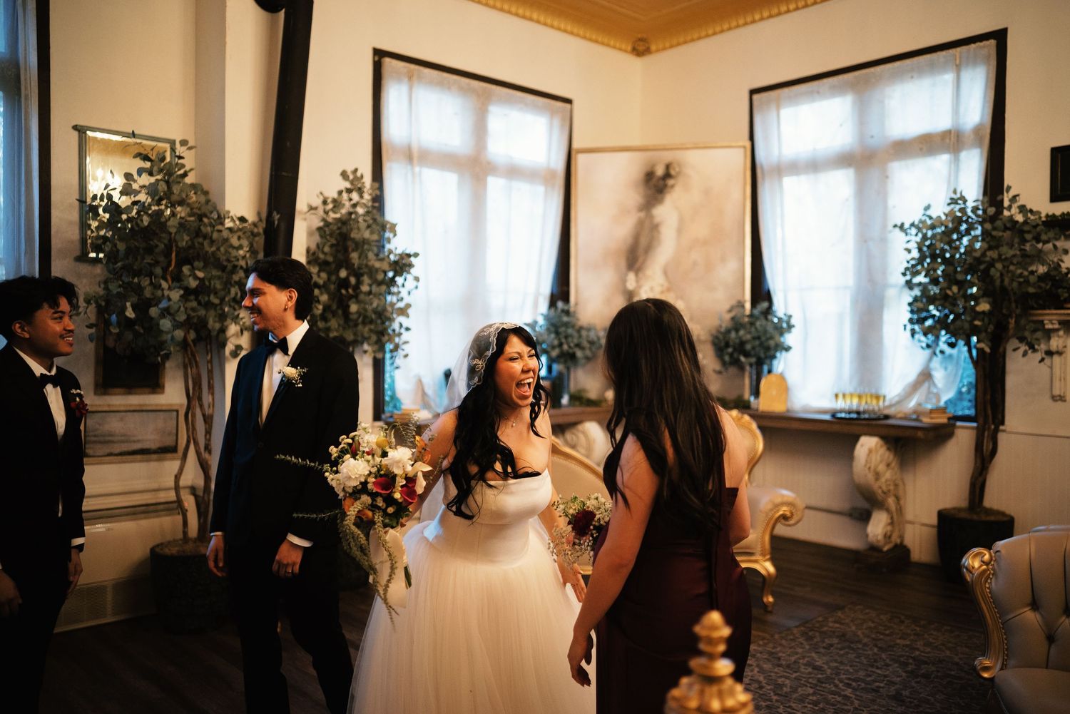Bride in tulle gown holding bouquet laughs with wedding party inside elegant venue after ceremony.