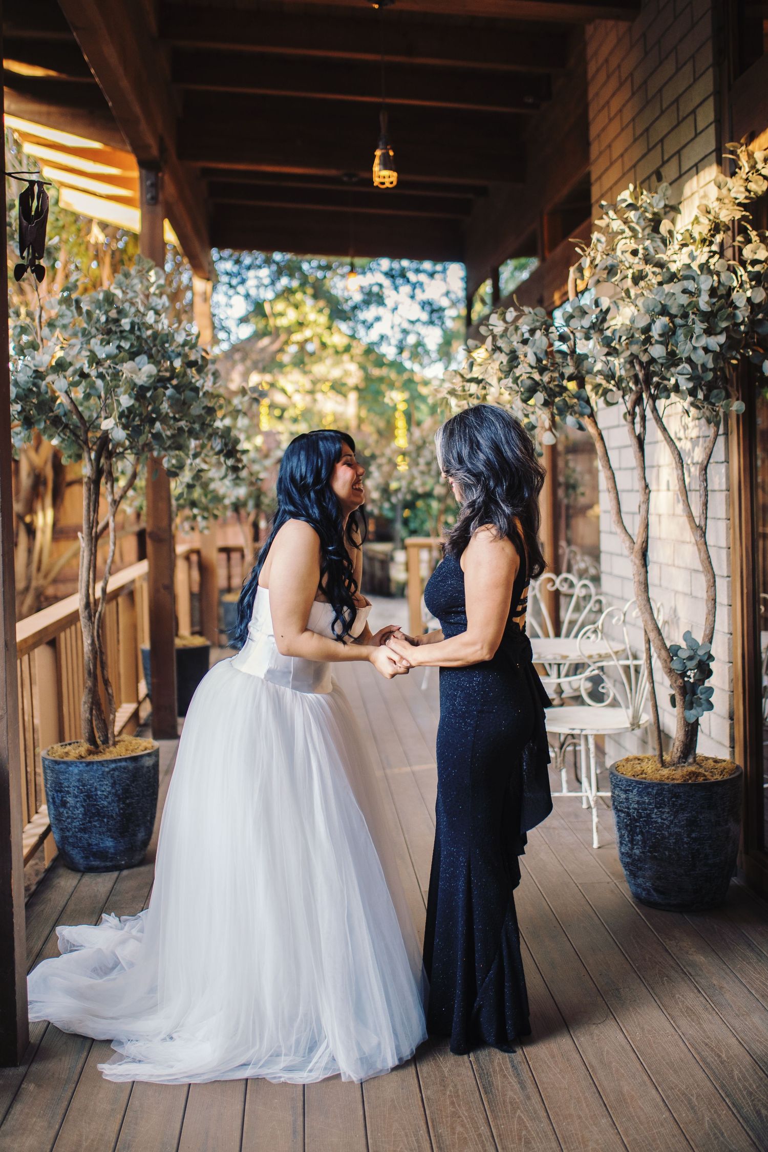 Bride in white tulle gown holds hands with her mother in a navy dress on a wooden porch.
