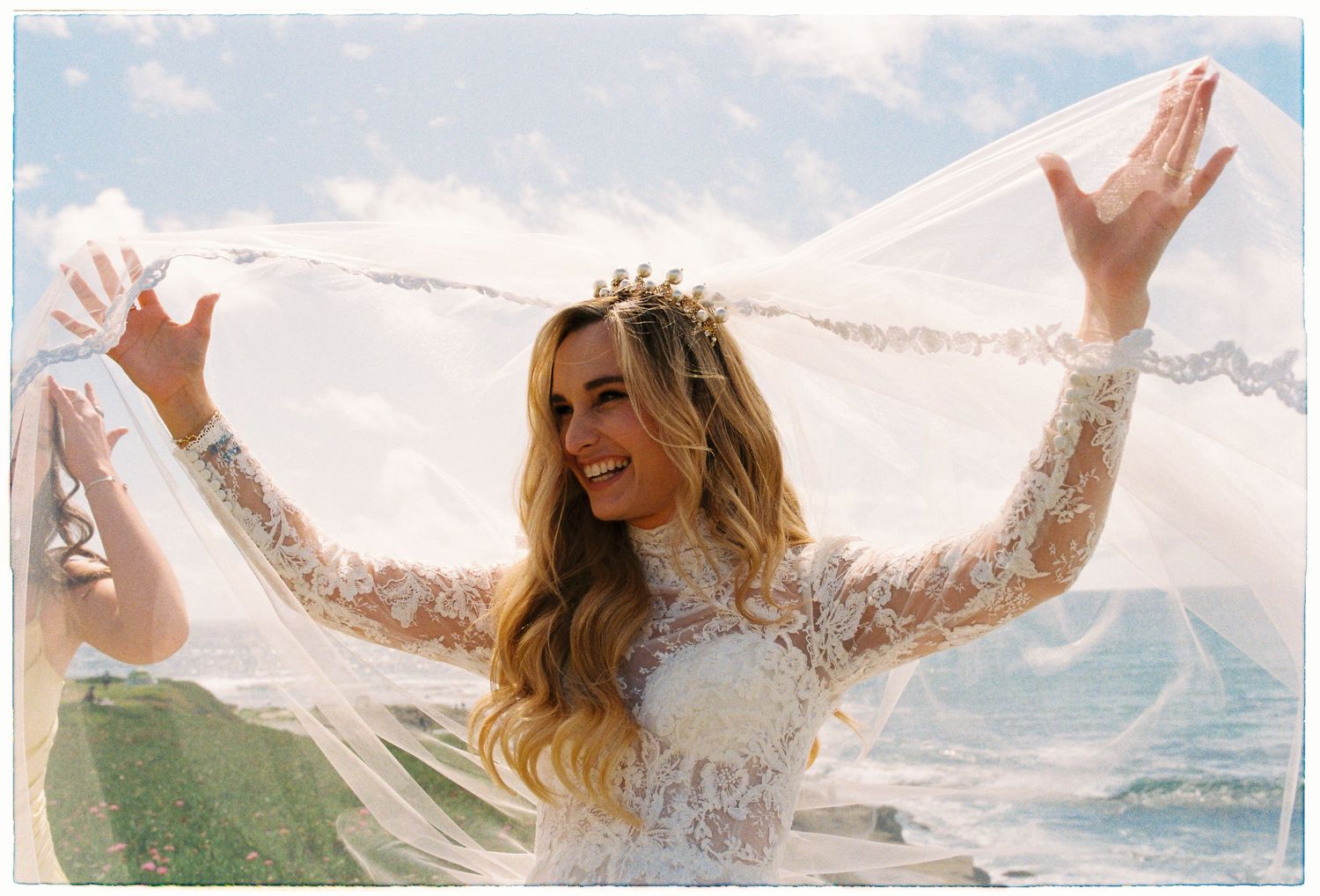 Smiling bride in lace gown holds her veil wide open against a bright blue ocean sky backdrop.