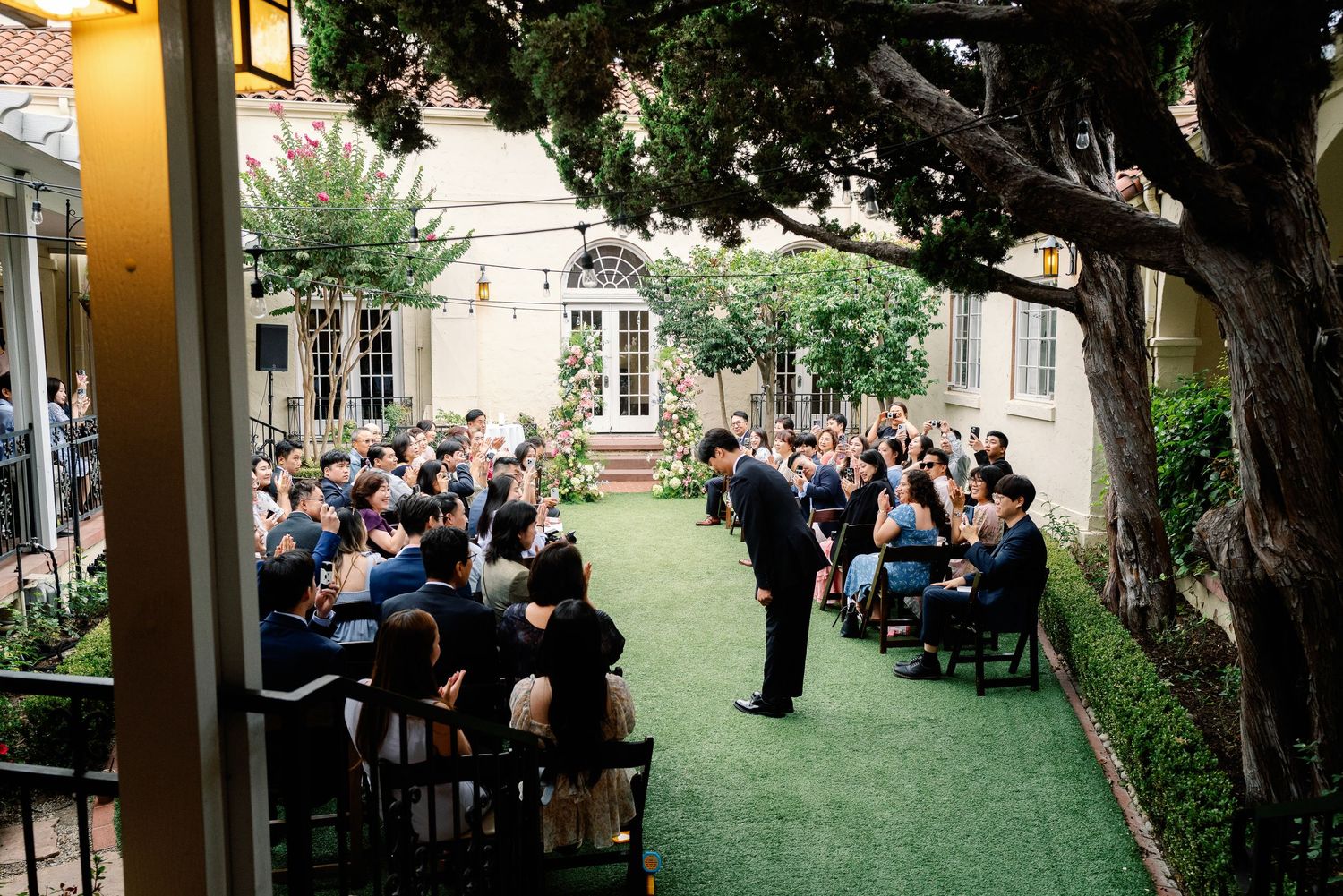 Groom bowing to seated wedding guests during an outdoor garden ceremony at a historic estate.