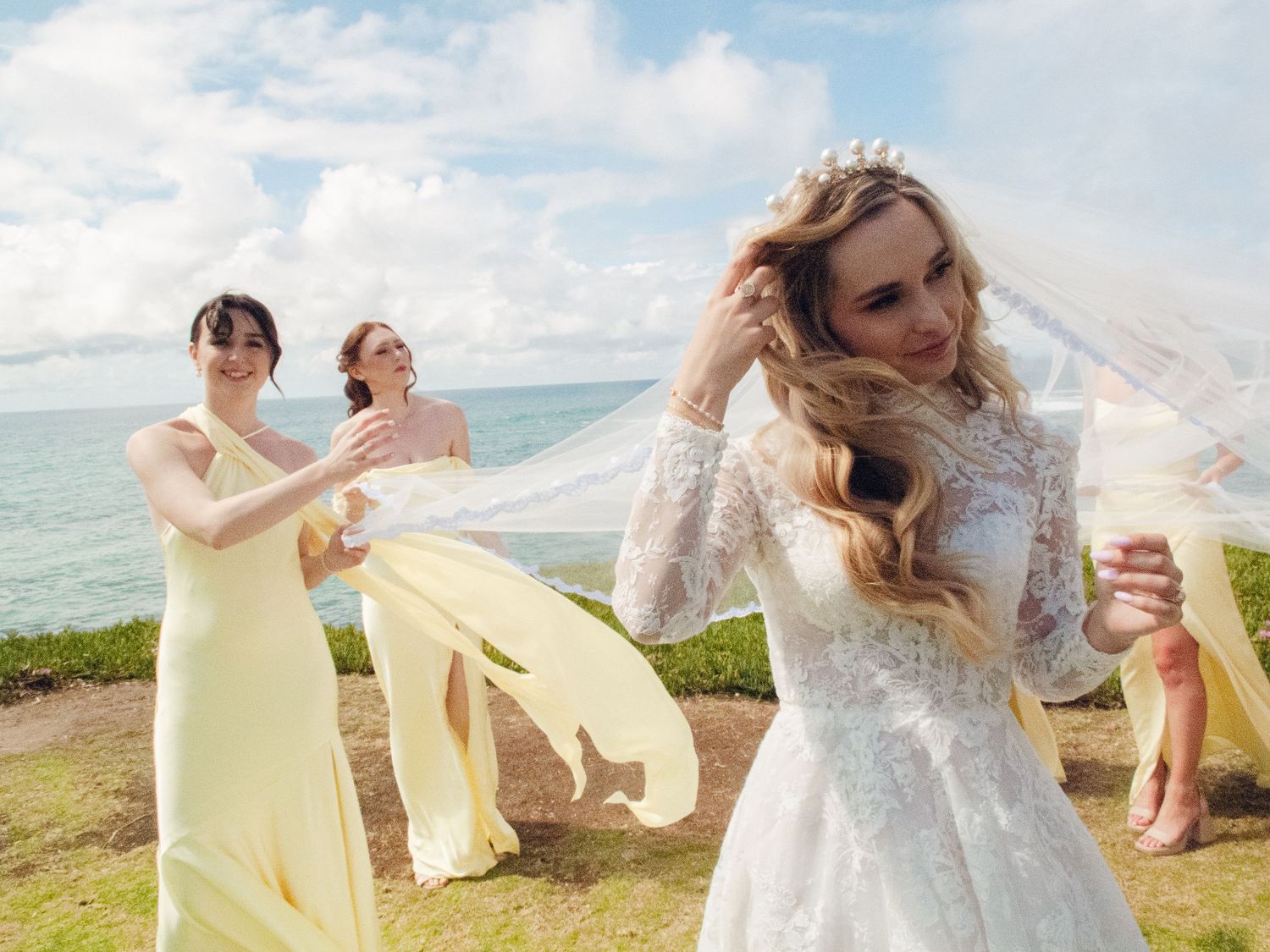 Bride in lace gown adjusts veil as bridesmaids in yellow dresses celebrate by the ocean.