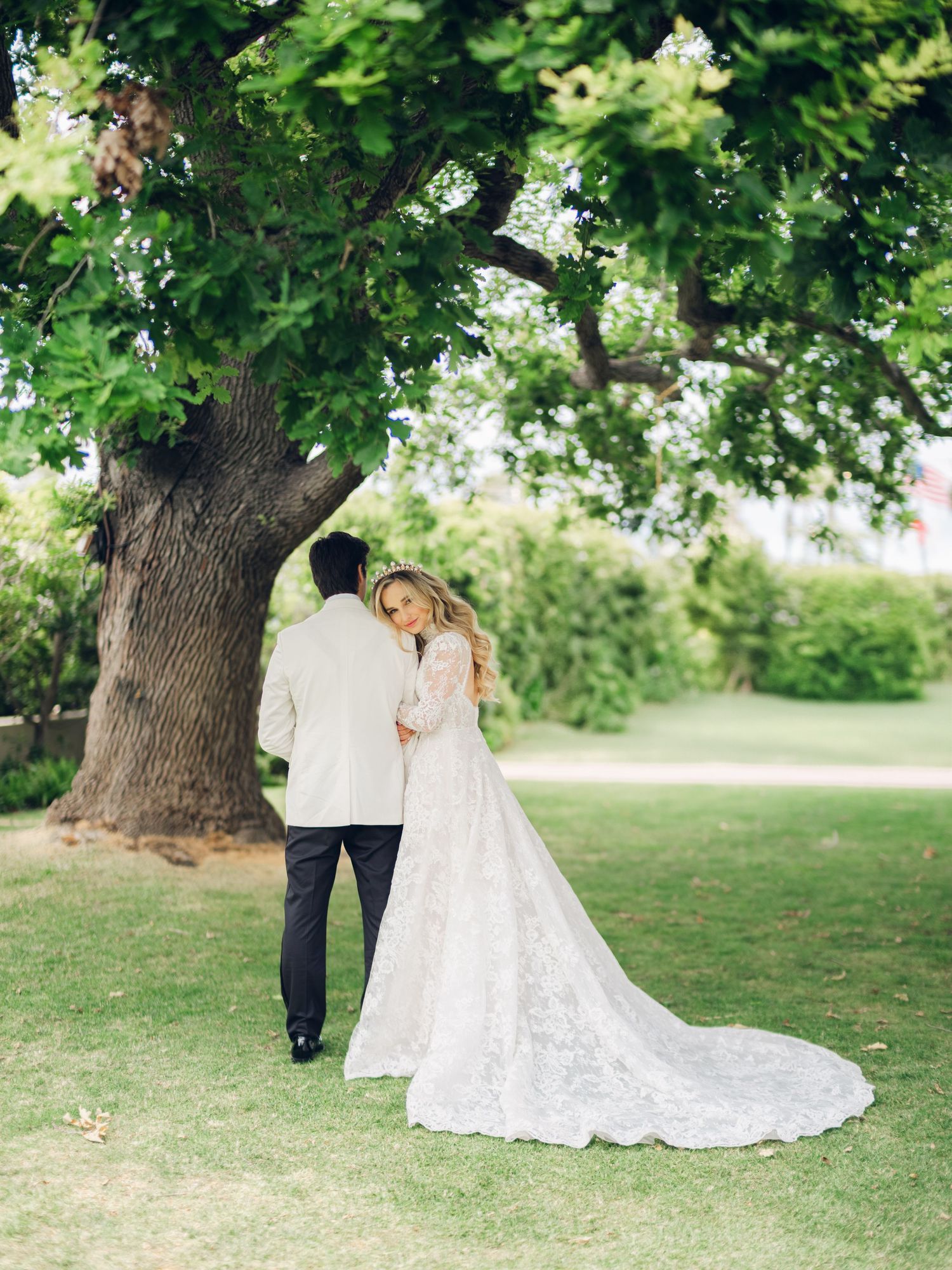 Bride in lace gown and groom in white tuxedo embrace under a large tree in a lush green garden.