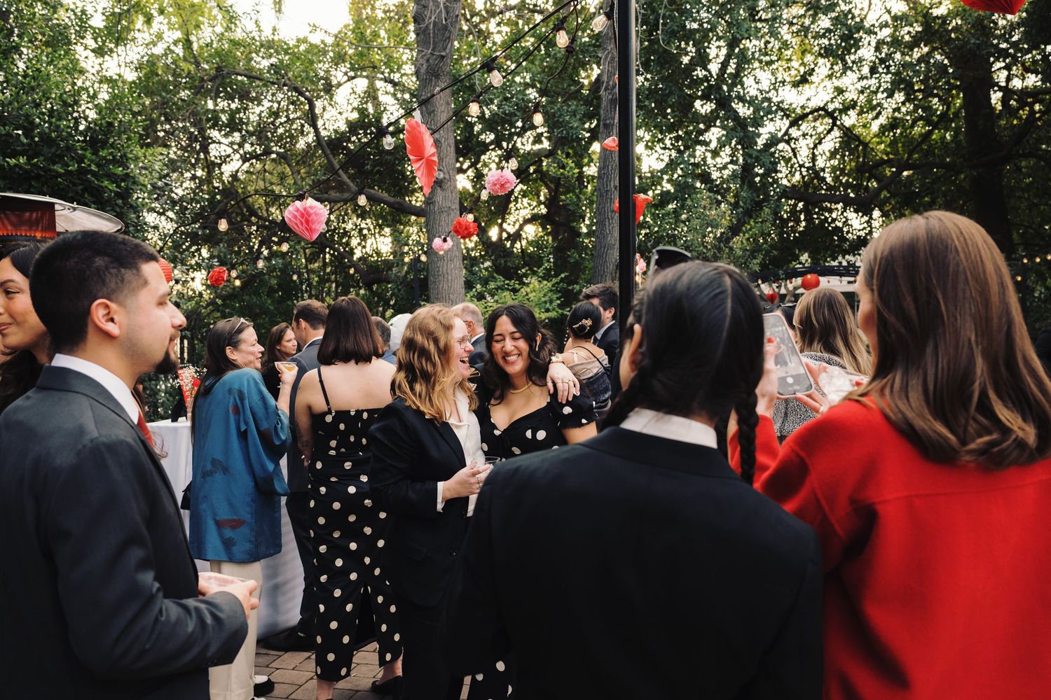 Guests mingling outdoors at a garden party with pink floral decorations hanging from trees.