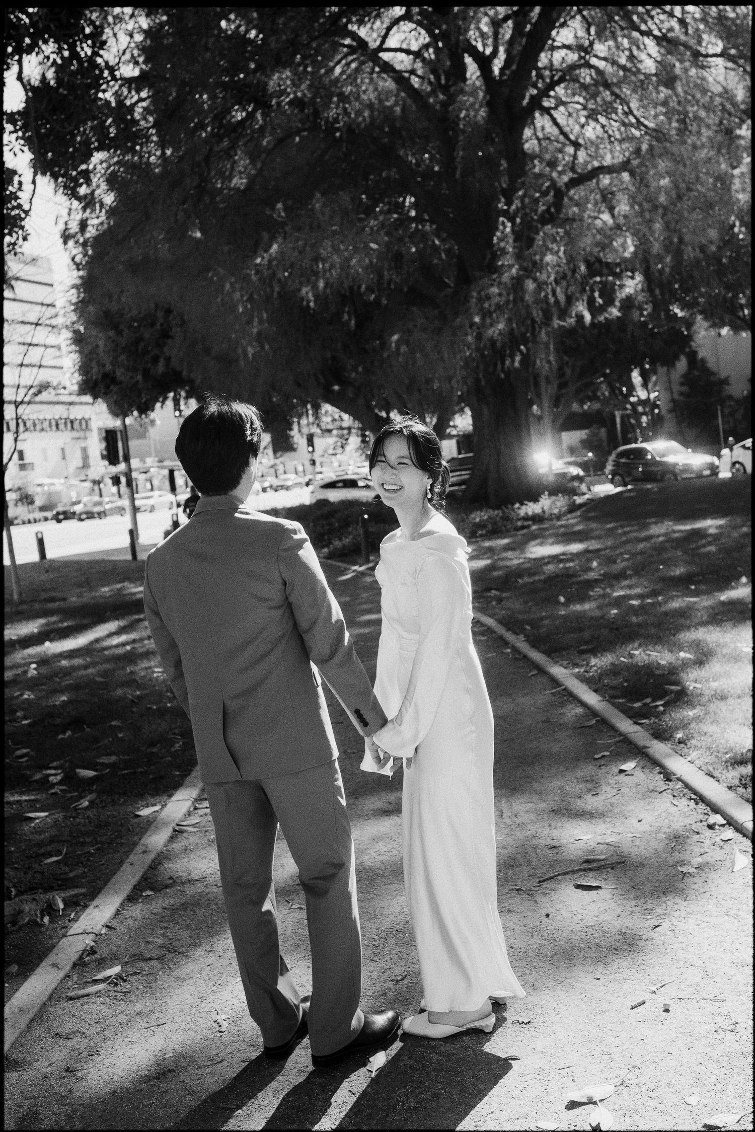 Black and white photo of a couple holding hands on a sunny path, bride in white gown smiling at groom in suit.