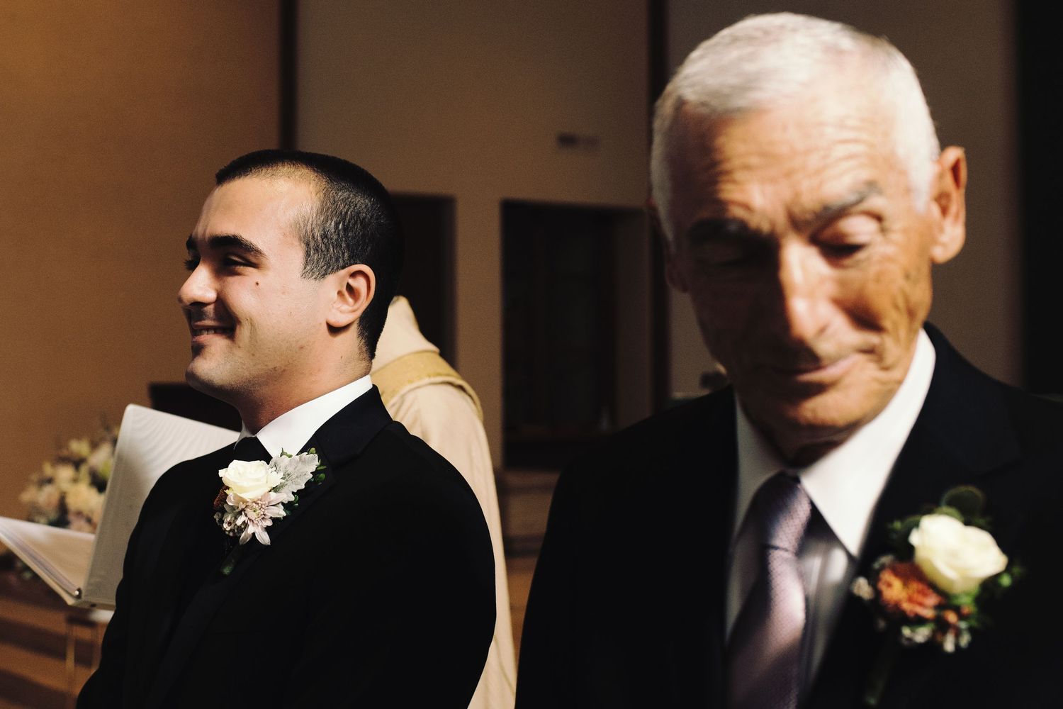 Two men in formal attire with boutonnieres stand together at a wedding ceremony indoors.