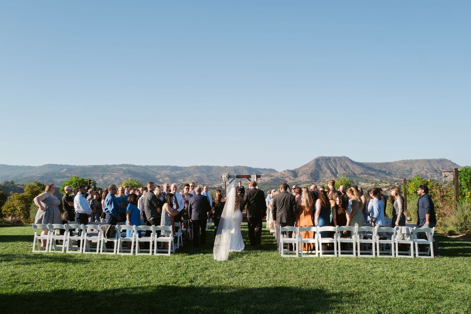 Wide shot of outdoor wedding ceremony on green lawn with stunning mountain range backdrop.