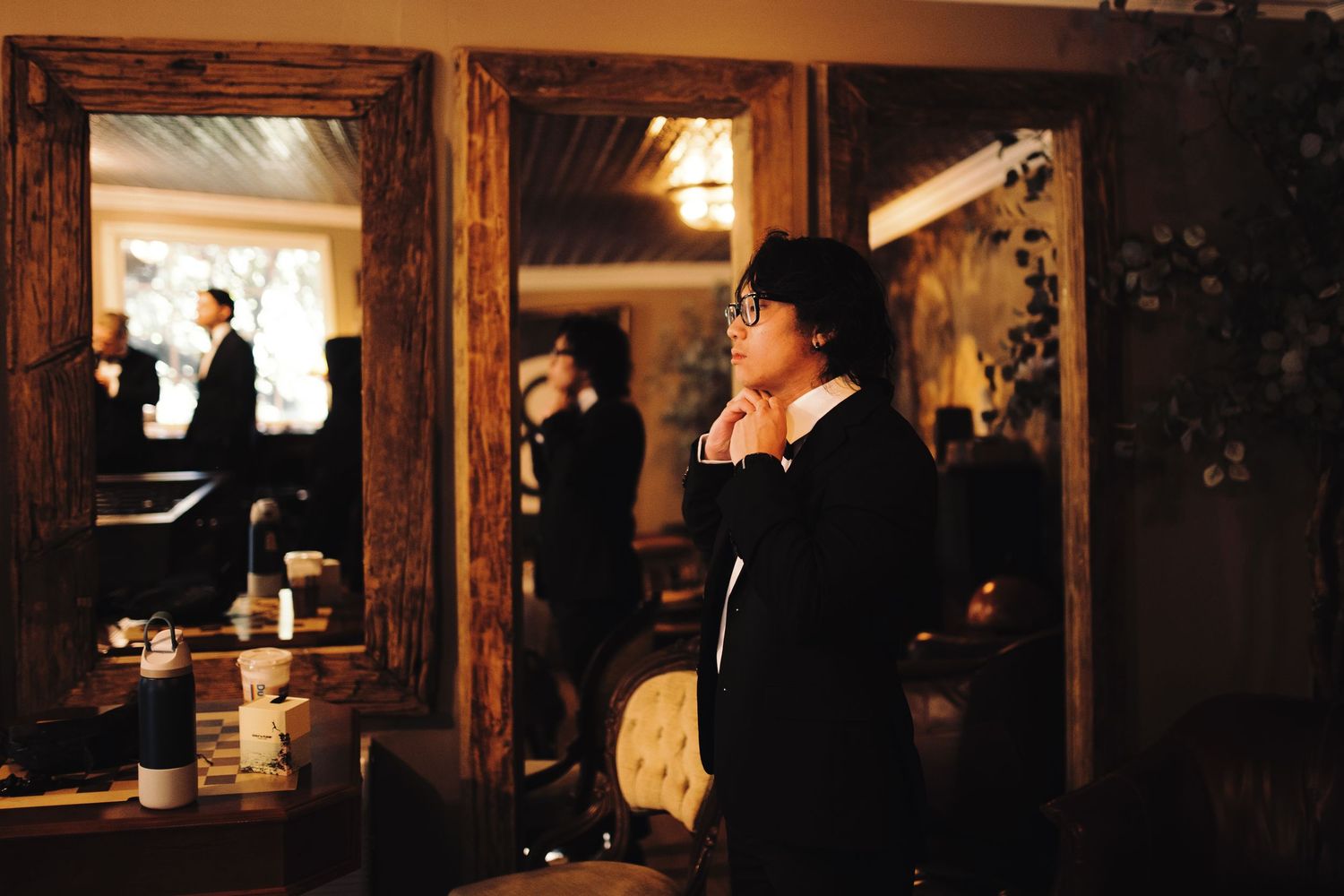 Groom in black tuxedo adjusts his collar while checking his reflection in a rustic framed mirror.
