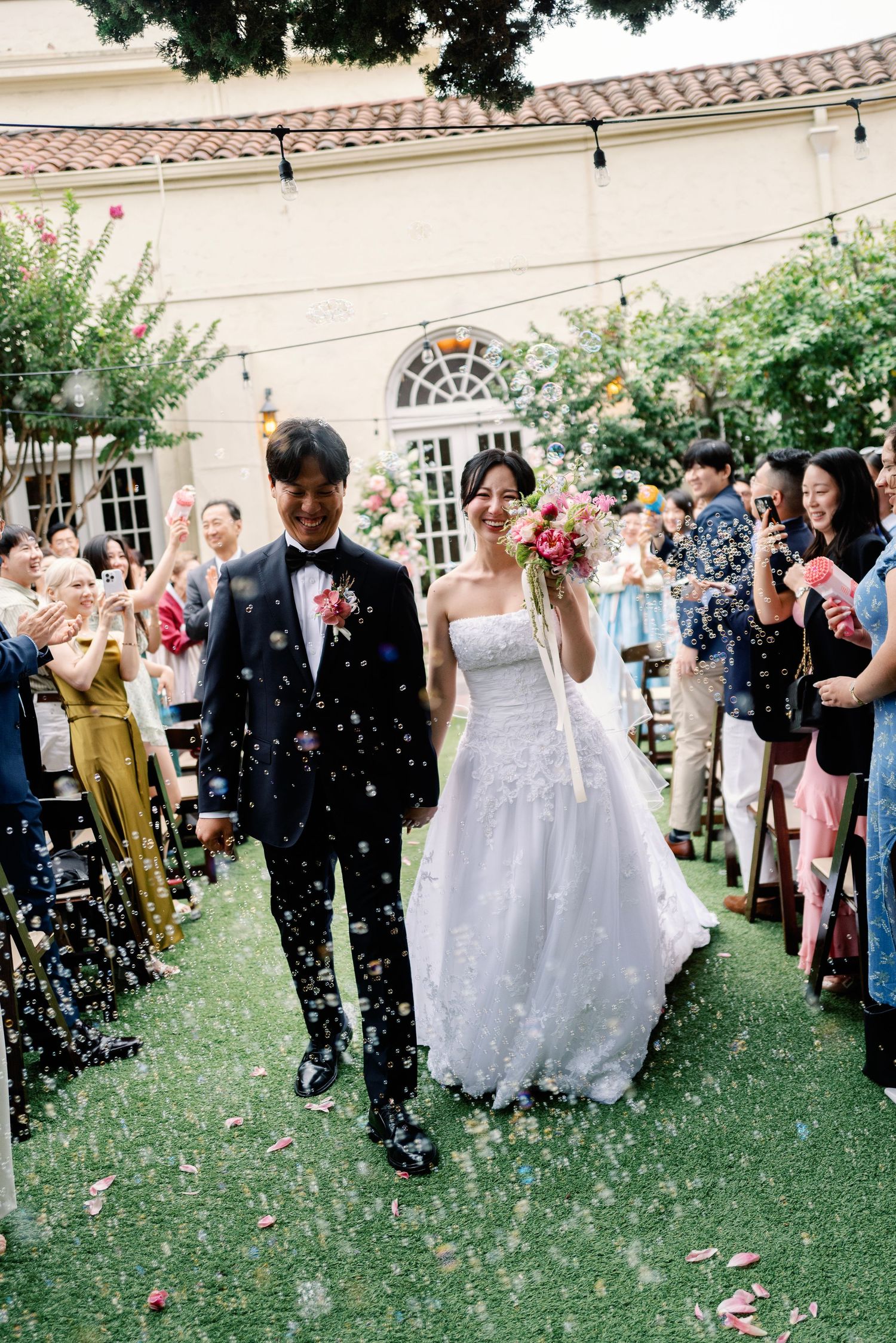 Smiling newlyweds walk down aisle through confetti shower at elegant outdoor garden wedding ceremony.