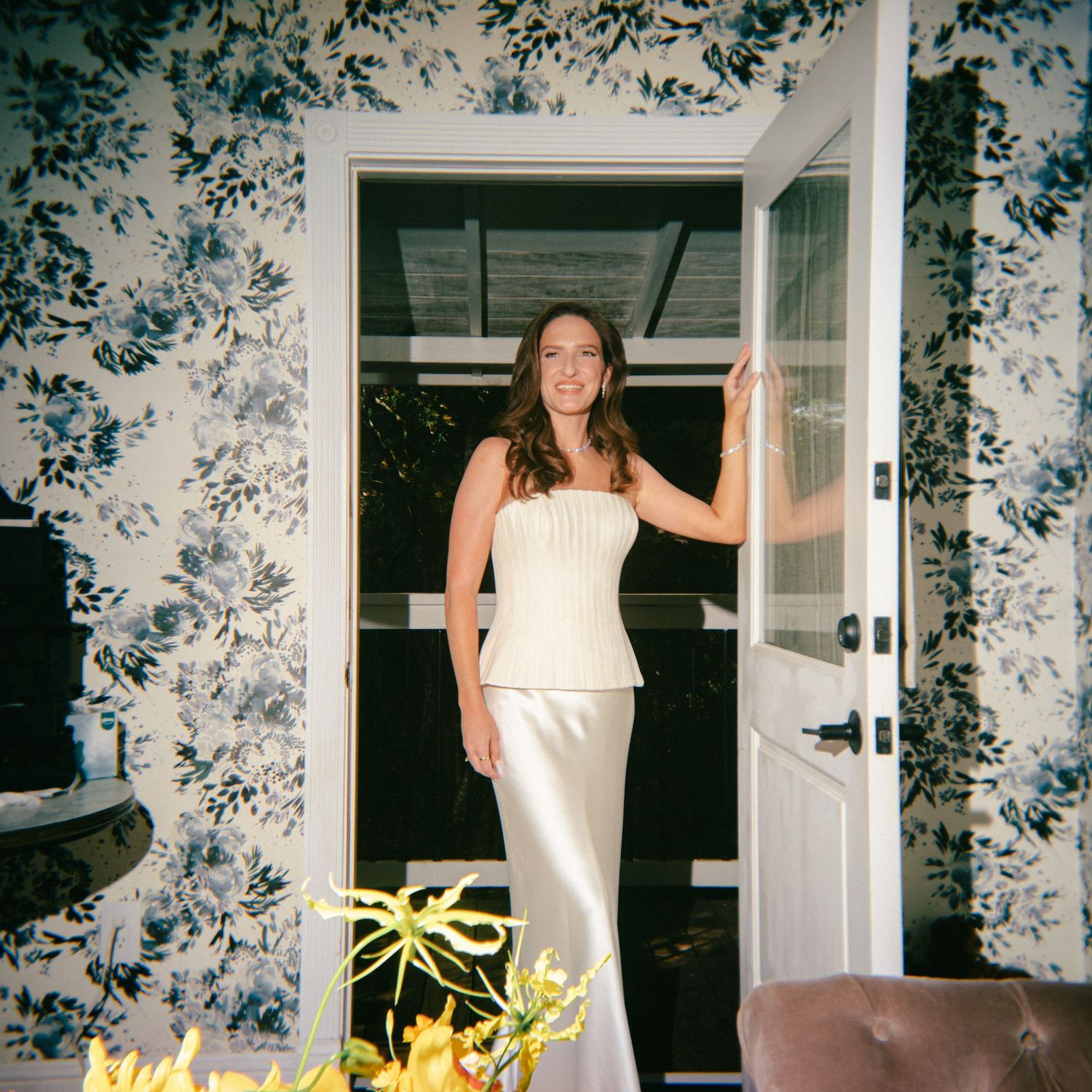 Woman in an elegant ivory satin bridal gown stands in a white doorway beside blue floral wallpaper, with yellow flowers in the foreground.