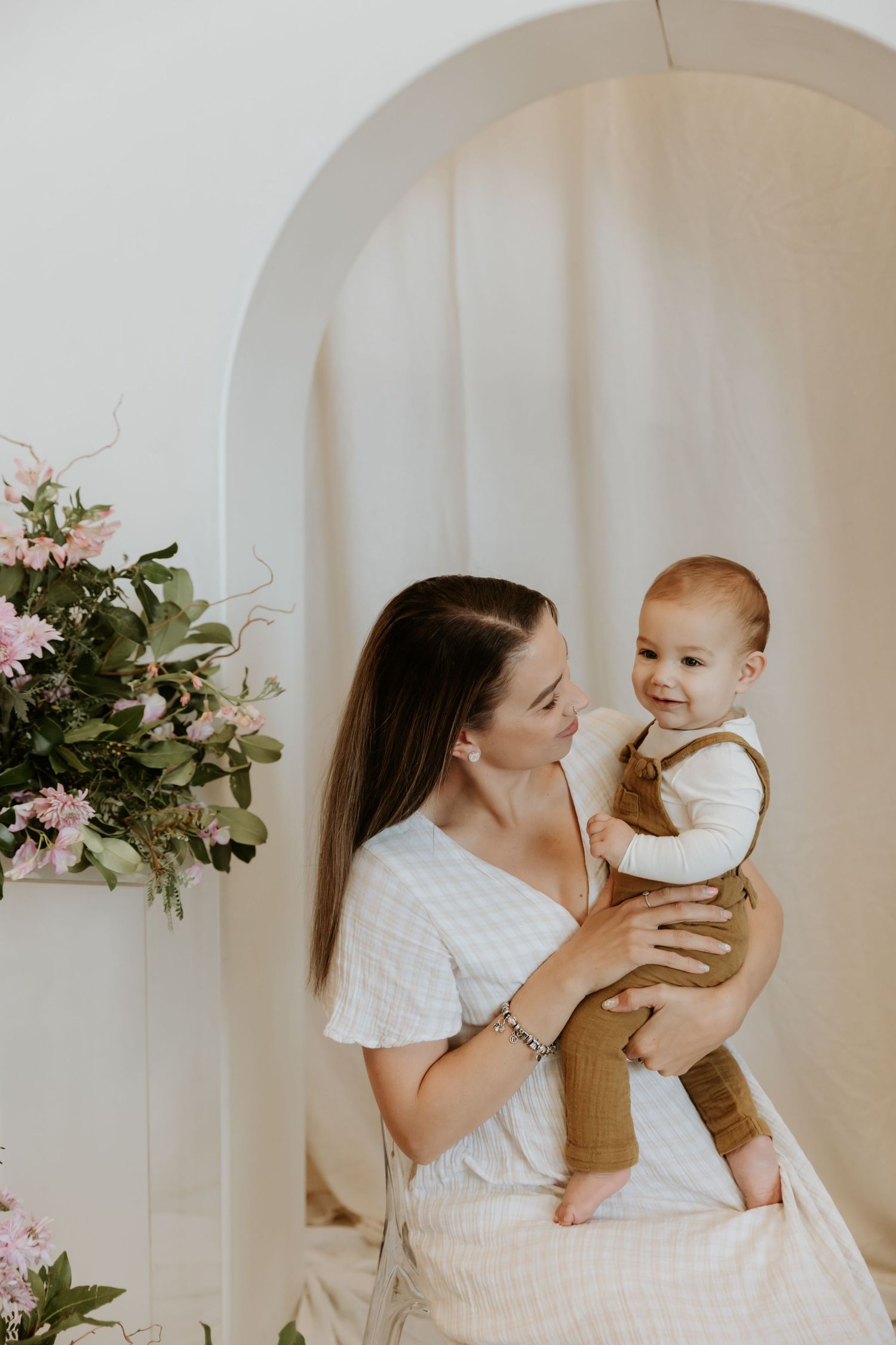 Mother in white dress playing with smiling baby against neutral backdrop with floral decor.
