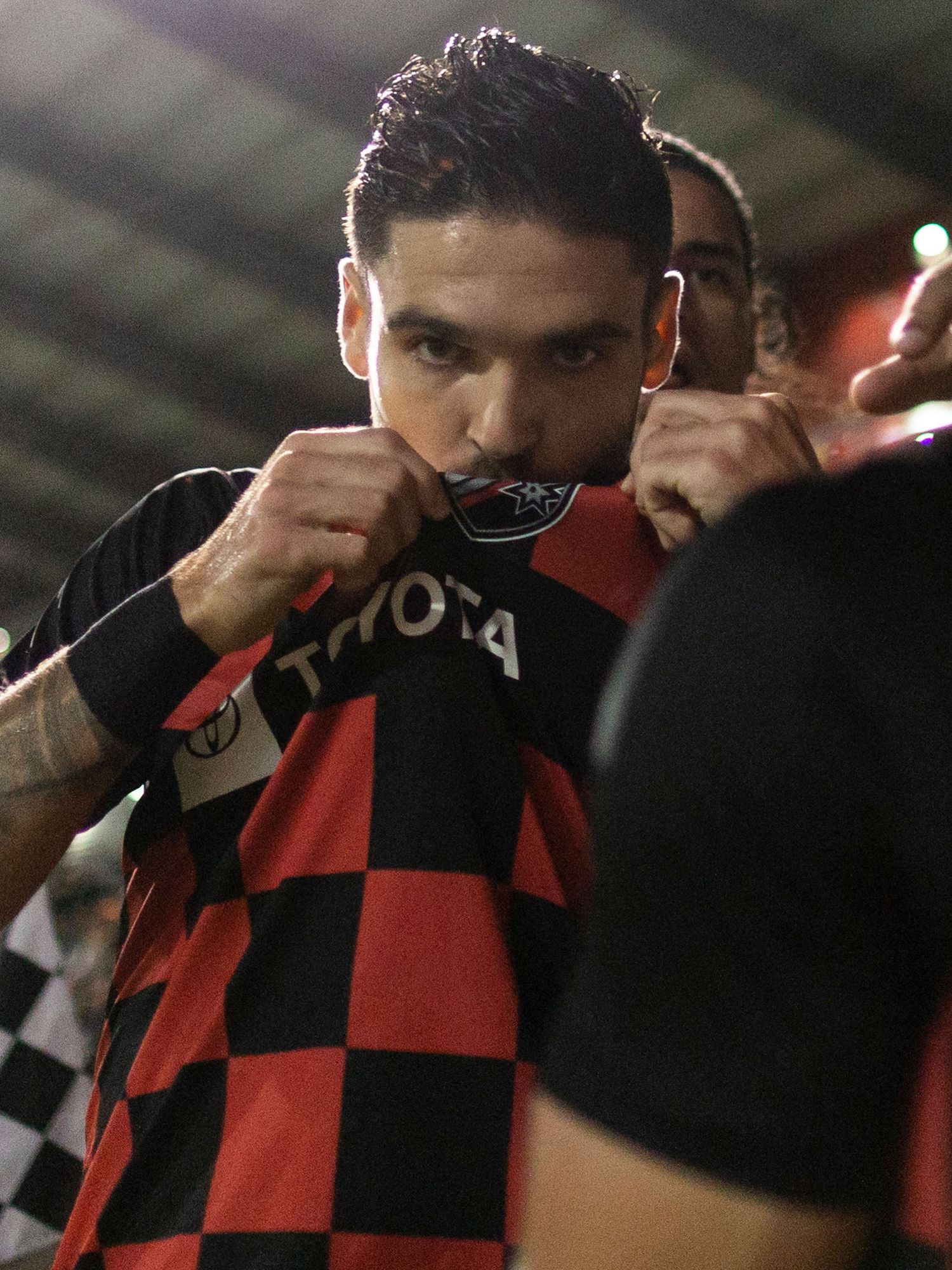 Soccer player in red and black checkered jersey kisses team badge after scoring goal during match.