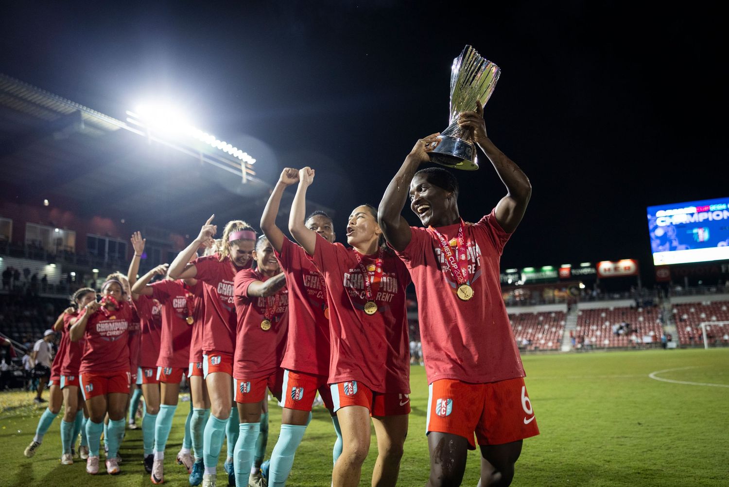Soccer team in red jerseys celebrates with trophy under stadium lights after winning championship match.