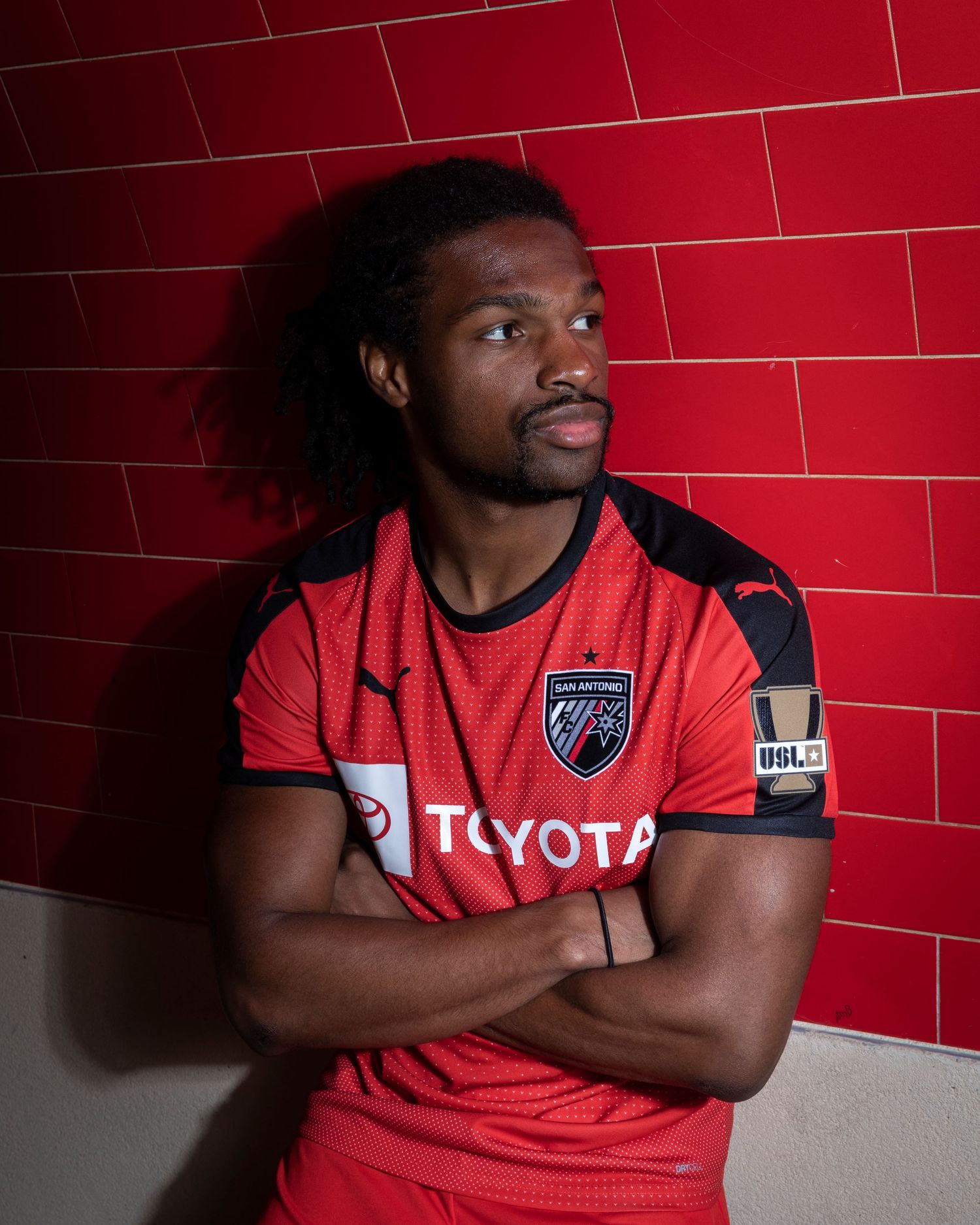 Athletic figure in red Toyota-sponsored jersey poses against red tile wall.