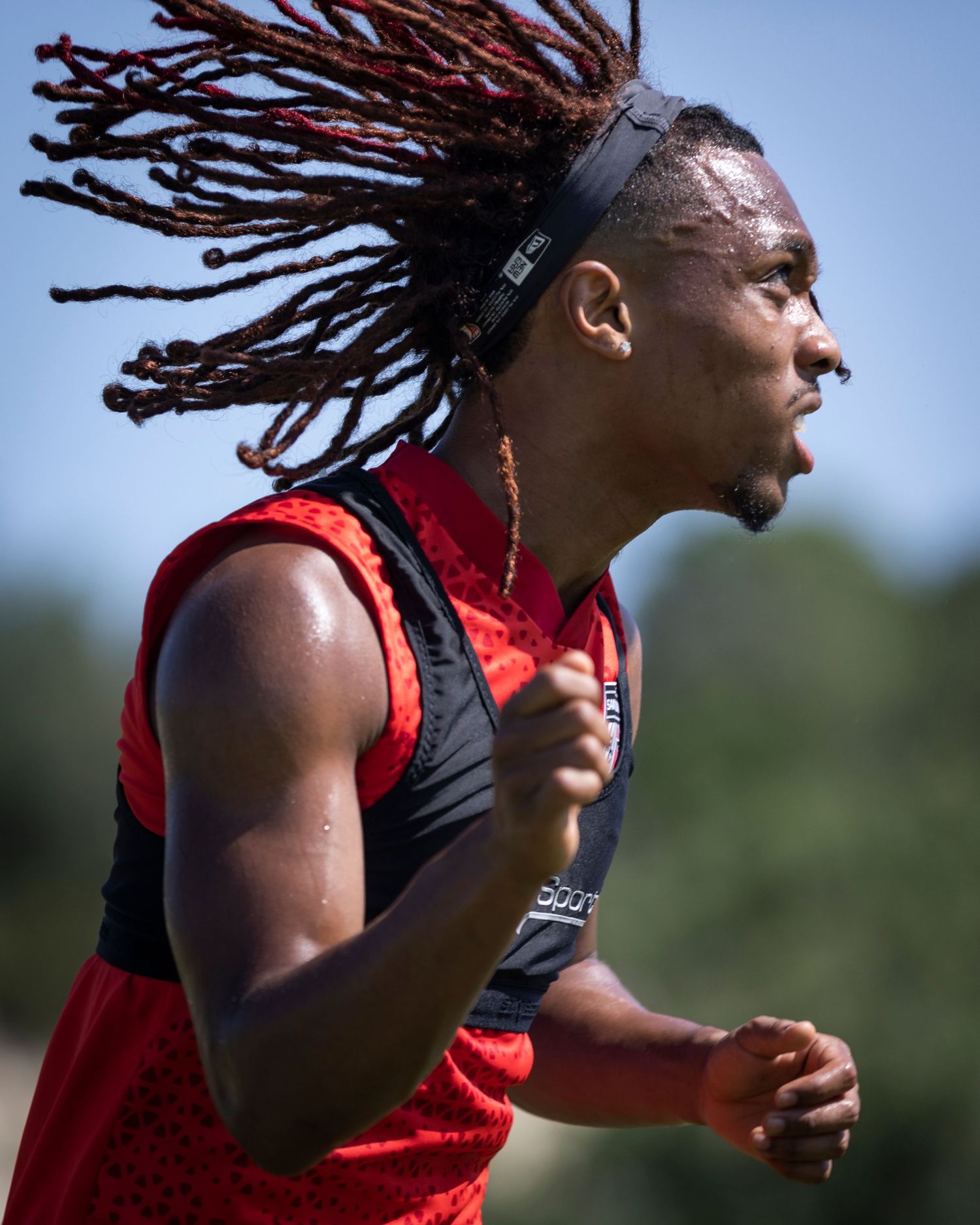 Athlete in red training vest running with dreadlocks flowing during practice session.