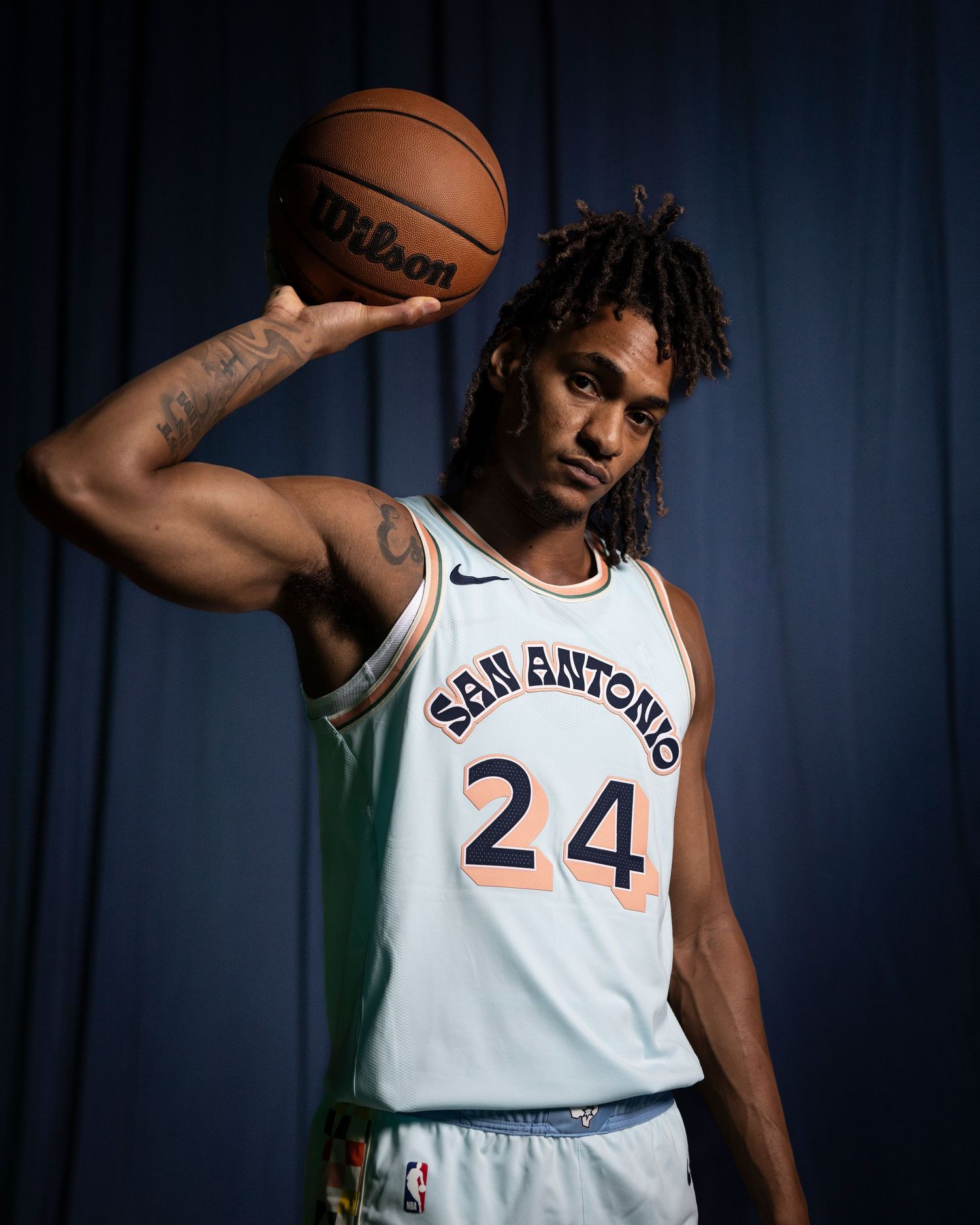 Player wearing San Antonio number 24 jersey holds basketball overhead in stylized portrait setting.