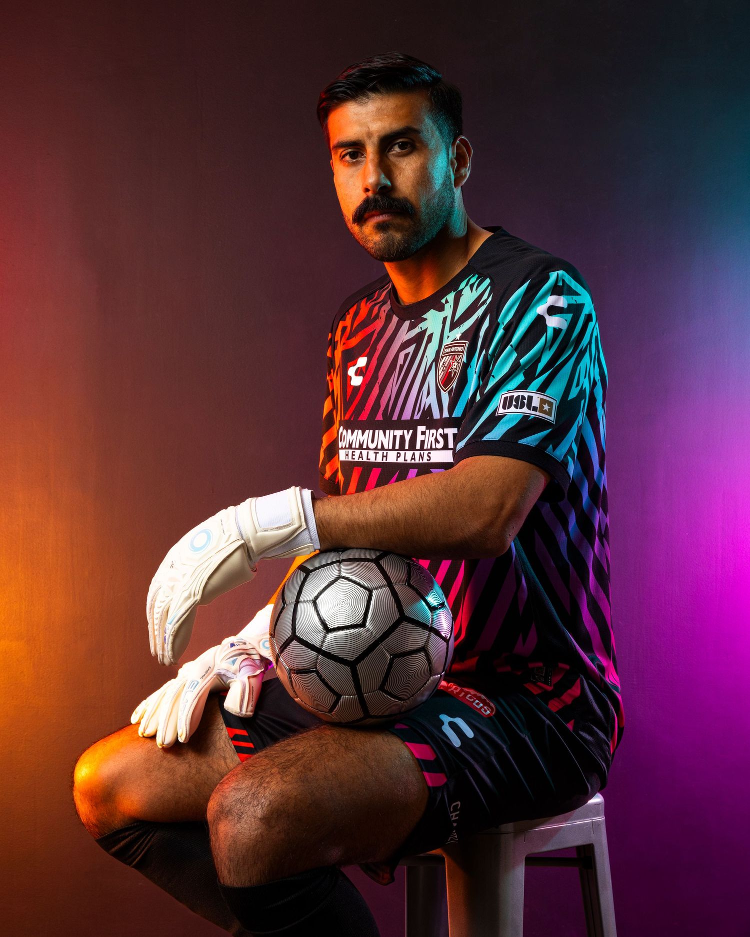 Goalkeeper in colorful jersey sits holding soccer ball against vibrant background.