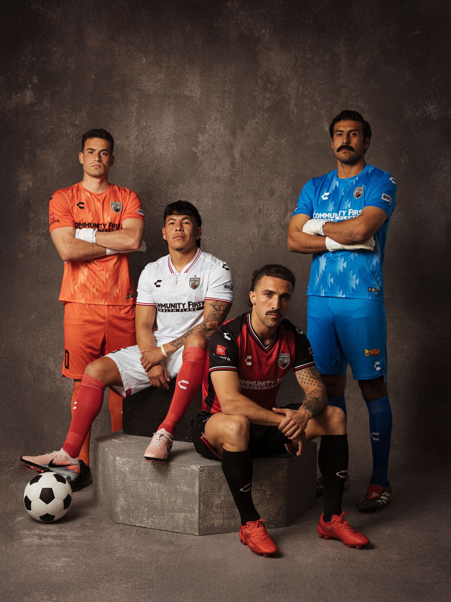 Four soccer players in colorful Chivas jerseys pose confidently in a studio, showcasing team uniforms in orange, white, red, and blue.