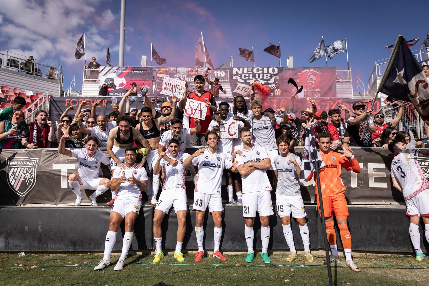 Soccer team celebrates victory with fans in stadium, players in white uniforms posing together with supporters behind them.