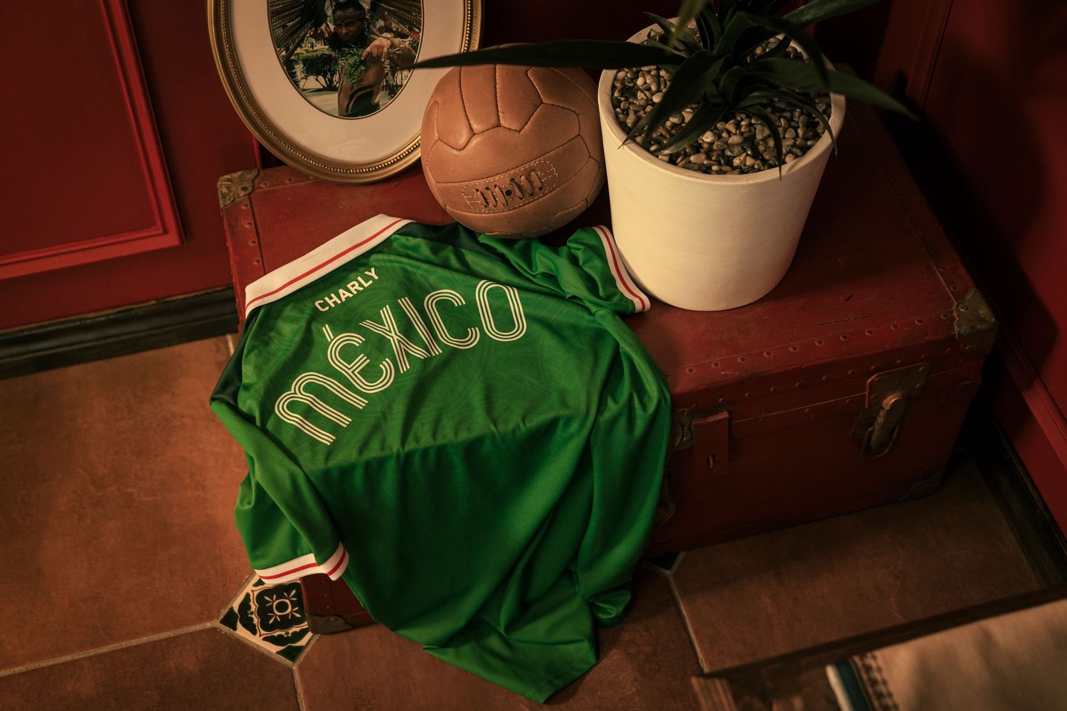 Green Mexico soccer jersey laid flat beside a vintage leather football and potted plant on a wooden trunk.