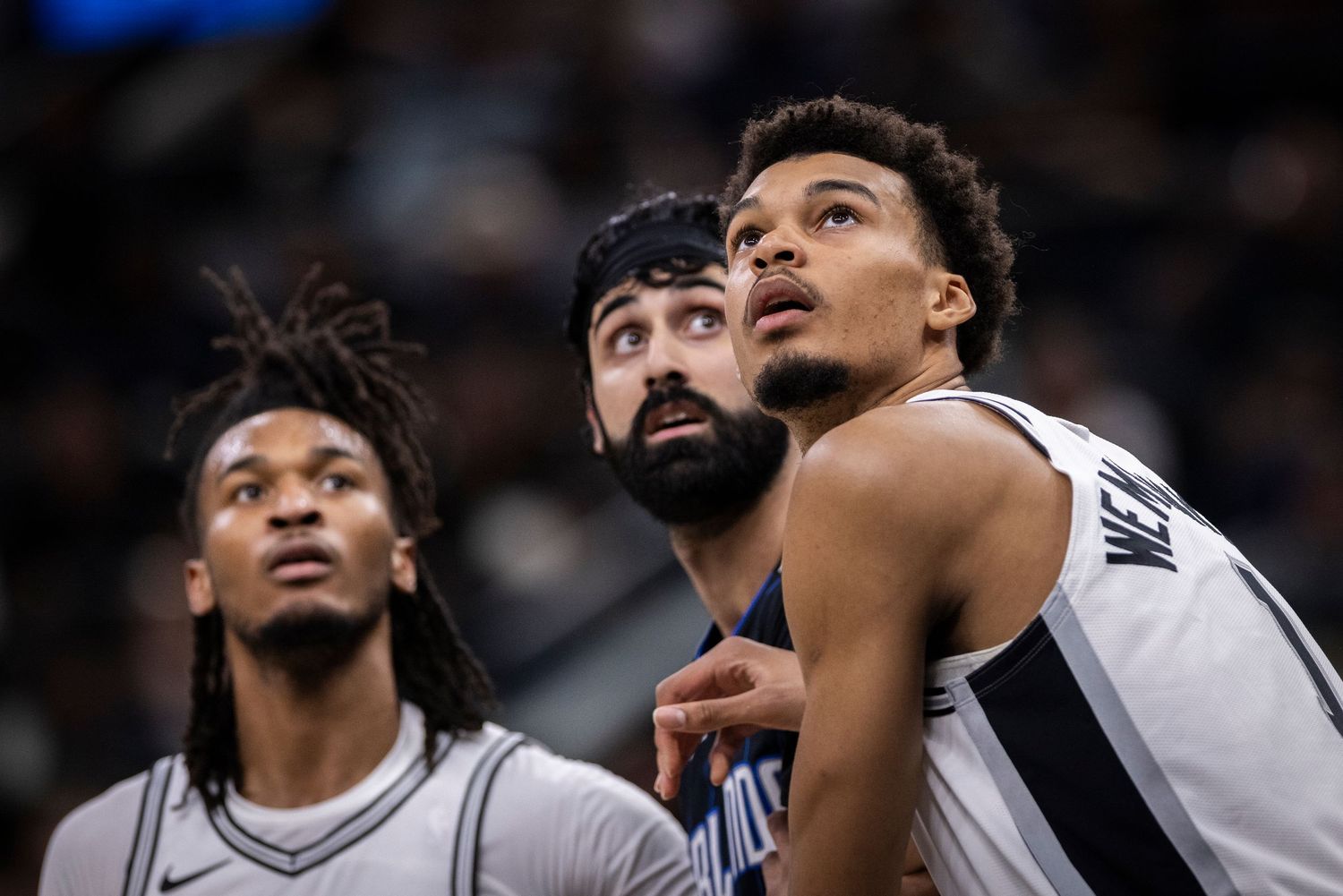 Three basketball players in black and white jerseys watching intently during a game. Victor Wembanyama
