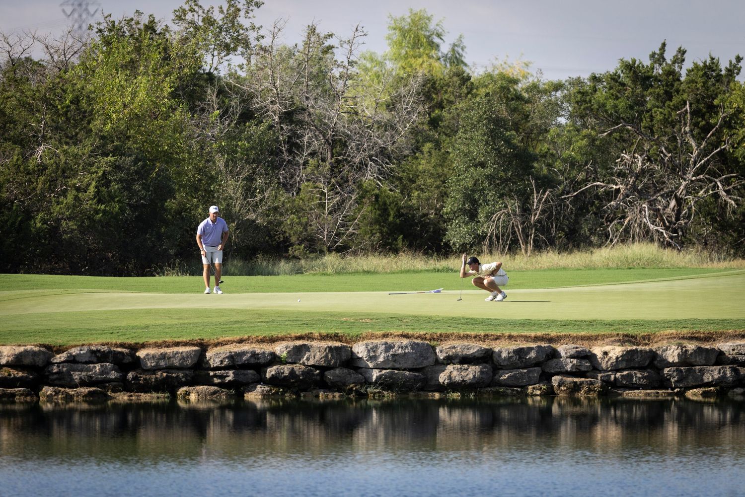 Golfers playing on scenic course with stone-lined water hazard and lush greenery.