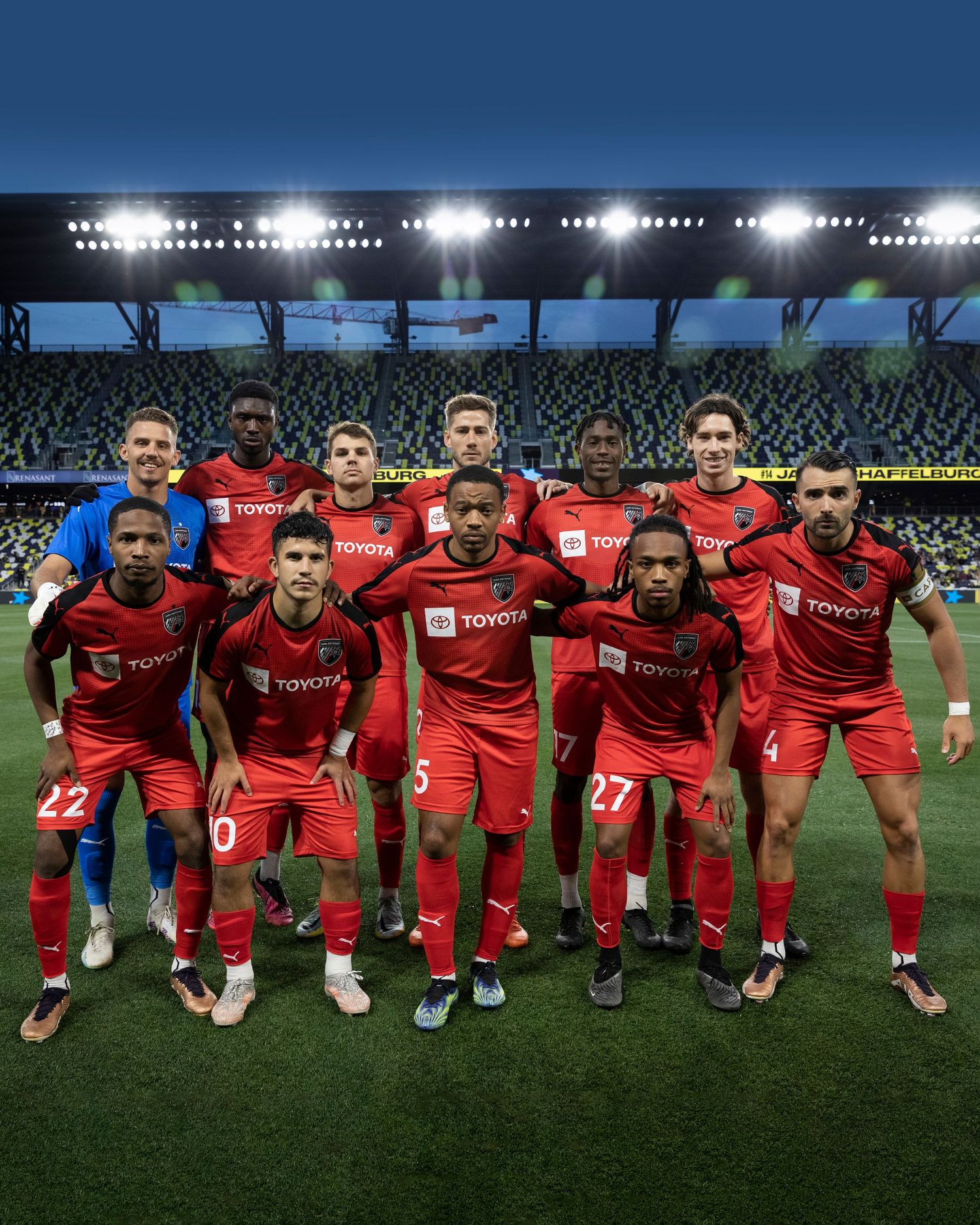 Soccer team in red uniforms poses for traditional pre-match lineup photo under stadium lights.