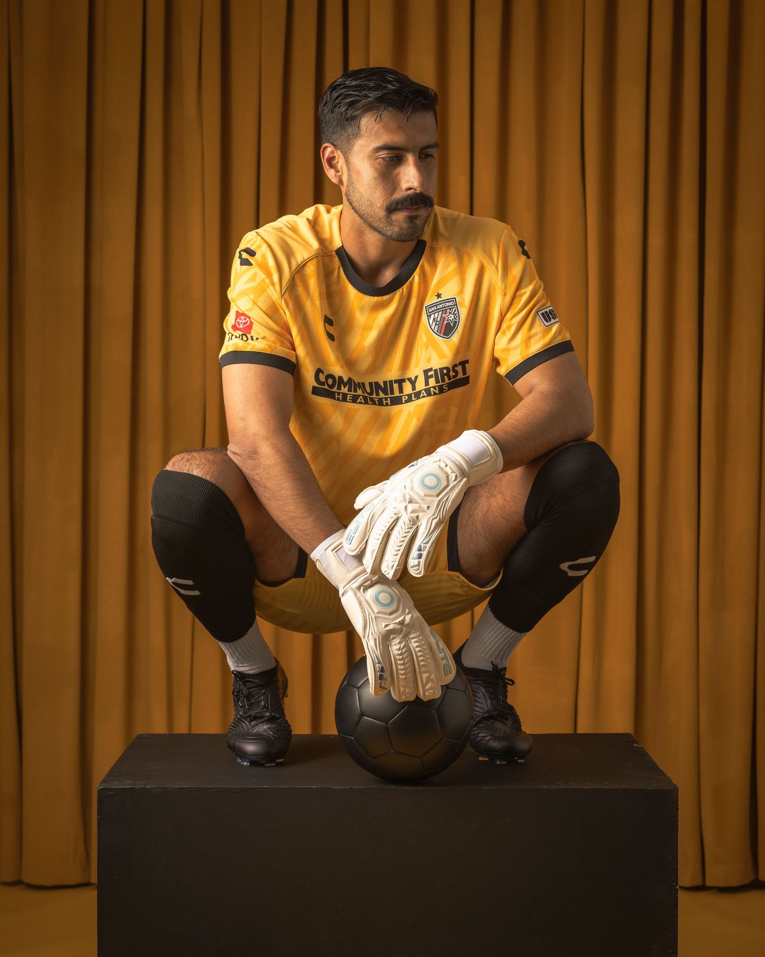 Goalkeeper in yellow uniform crouches on black platform against gold curtain backdrop.