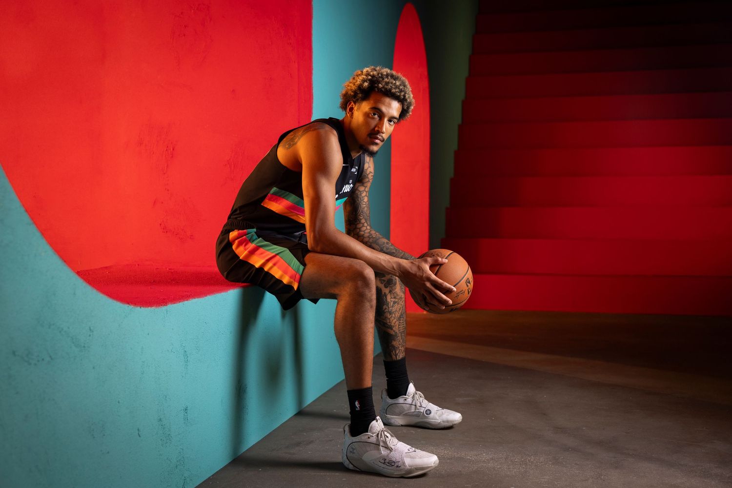 NBA basketball player in black and orange uniform sits with a ball against a vibrant red and teal backdrop.