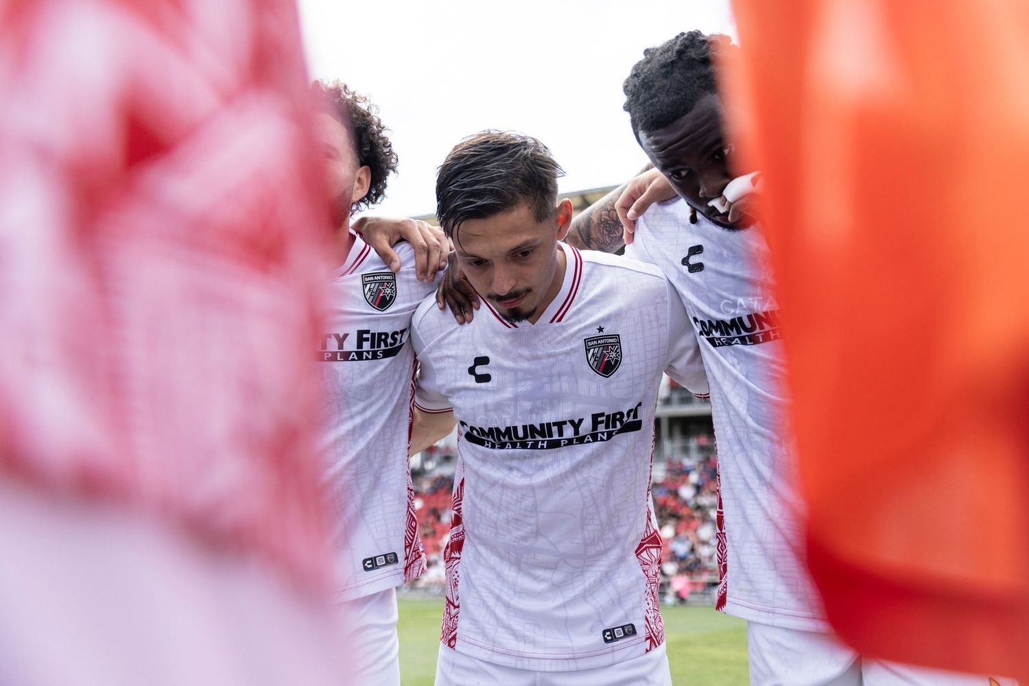 Soccer players huddling together in white uniforms before a match at a red-seated stadium.