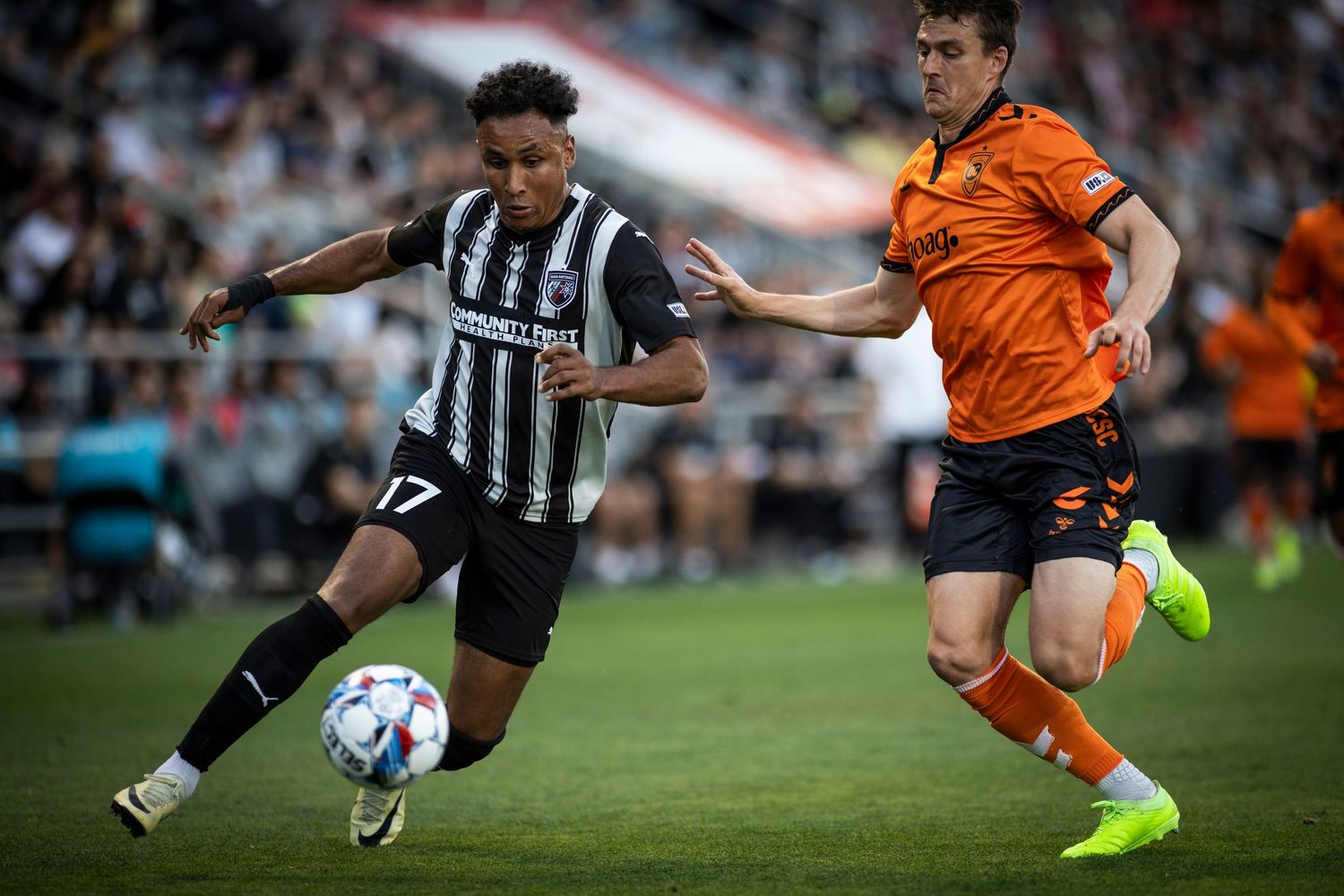 Two soccer players compete for the ball during a match between teams in black-white stripes and orange.