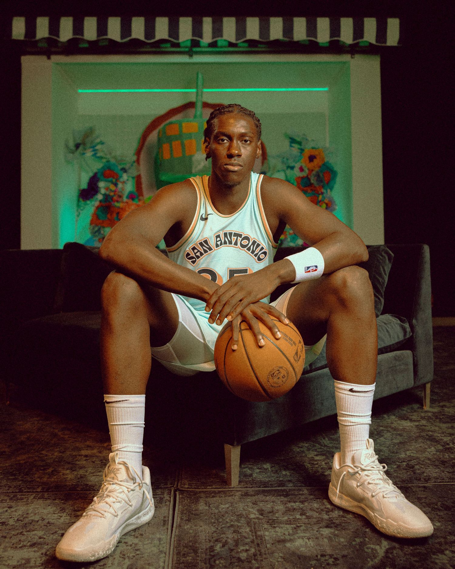 Athlete in San Antonio uniform sits holding basketball against artistic backdrop.
