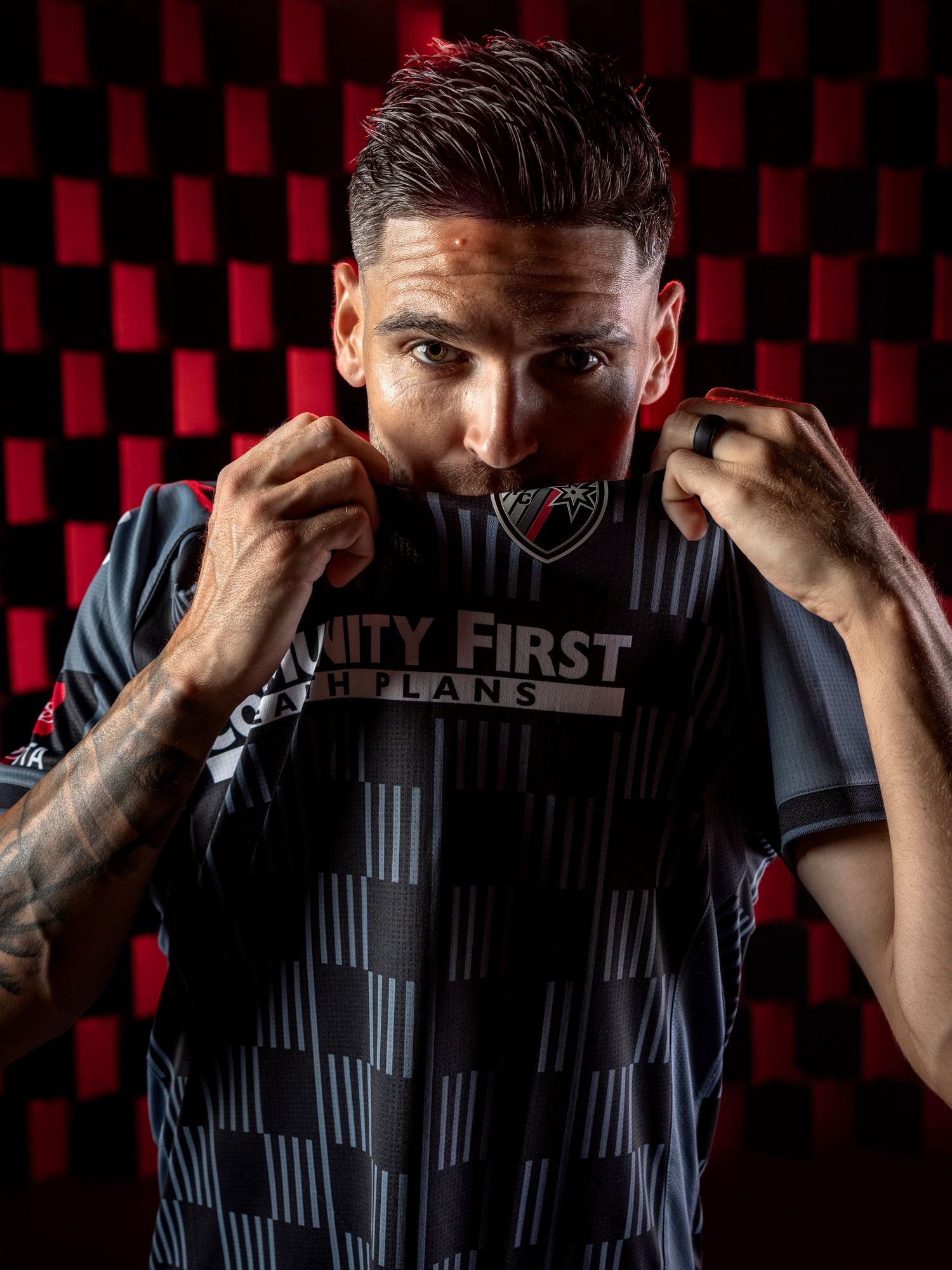Athletic figure in a dark striped jersey with 'UNITY FIRST' text poses against red and black checkered background.