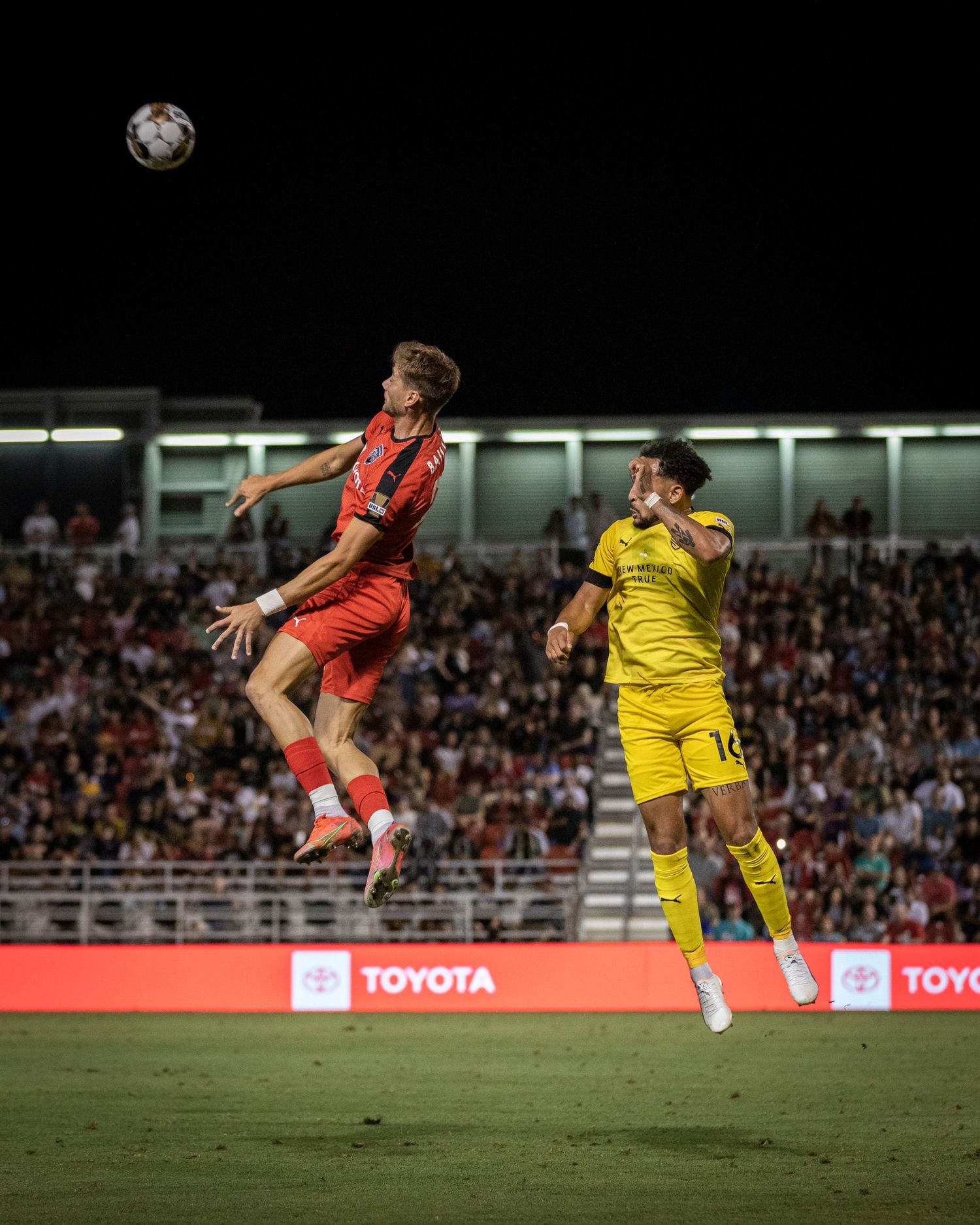 Soccer player in red jersey leaps high to head the ball during a match against yellow-uniformed opponents.
