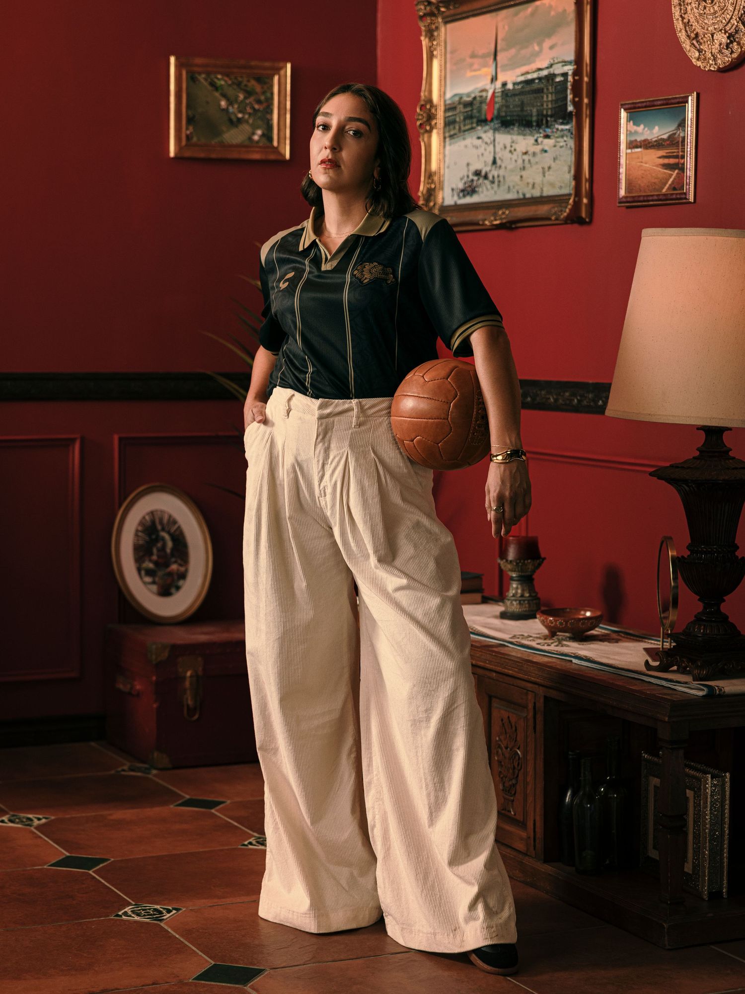 Woman in vintage soccer jersey and wide-leg trousers holding a retro leather football in a red-walled room.