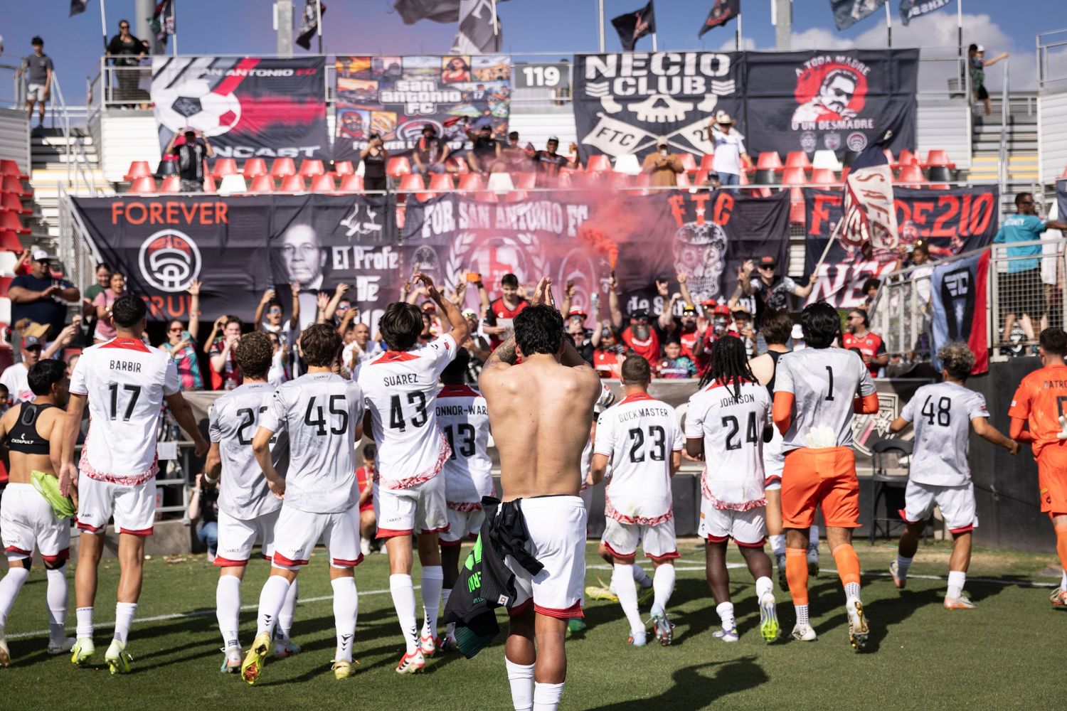 San Antonio FC players celebrate with passionate fans holding banners and red smoke flares after a soccer victory.